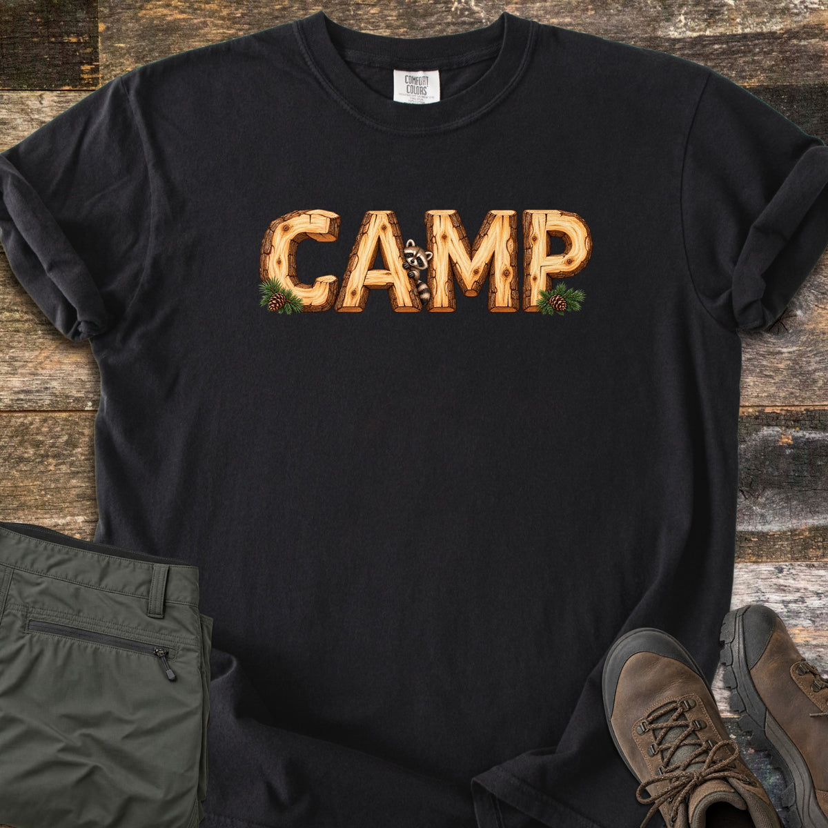 Camp Comfort Colors T-shirt in Black, laid flat. The word CAMP is printed in letters made of cut wooden logs, with a raccoon peeking out between the A and M and pine sprigs with pinecones at each end. Olive pants and brown hiking boots sit on weathered wood planks.