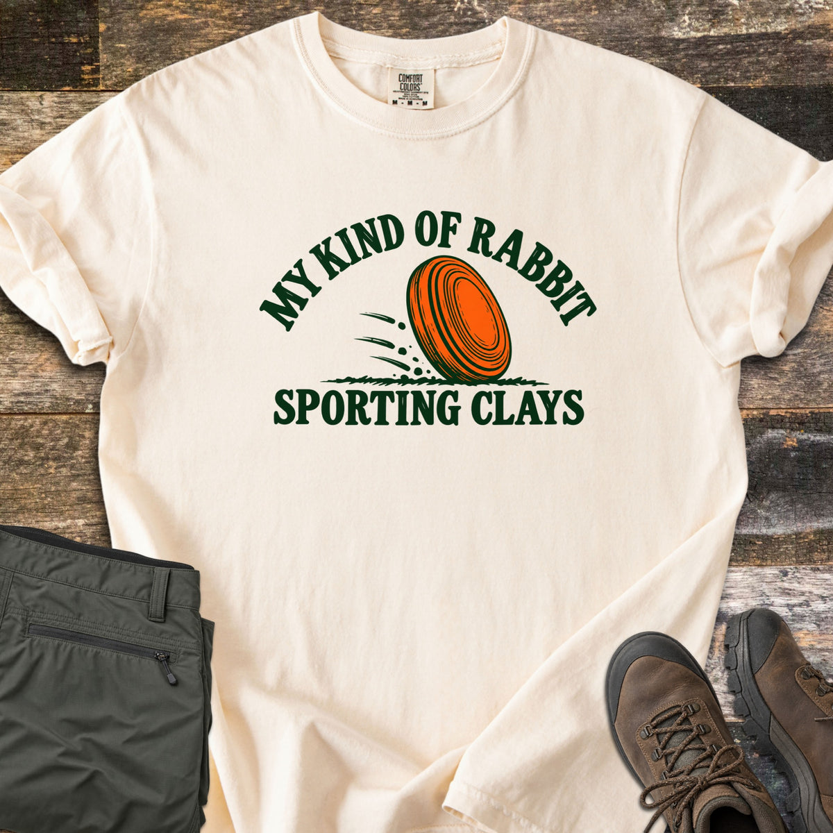 My Kind of Rabbit Comfort Colors T-shirt in Ivory, laid flat. An orange clay target rolls through grass between the curved words My Kind of Rabbit and Sporting Clays in dark green. Olive hiking pants and brown boots lie beside it on weathered wood planks.