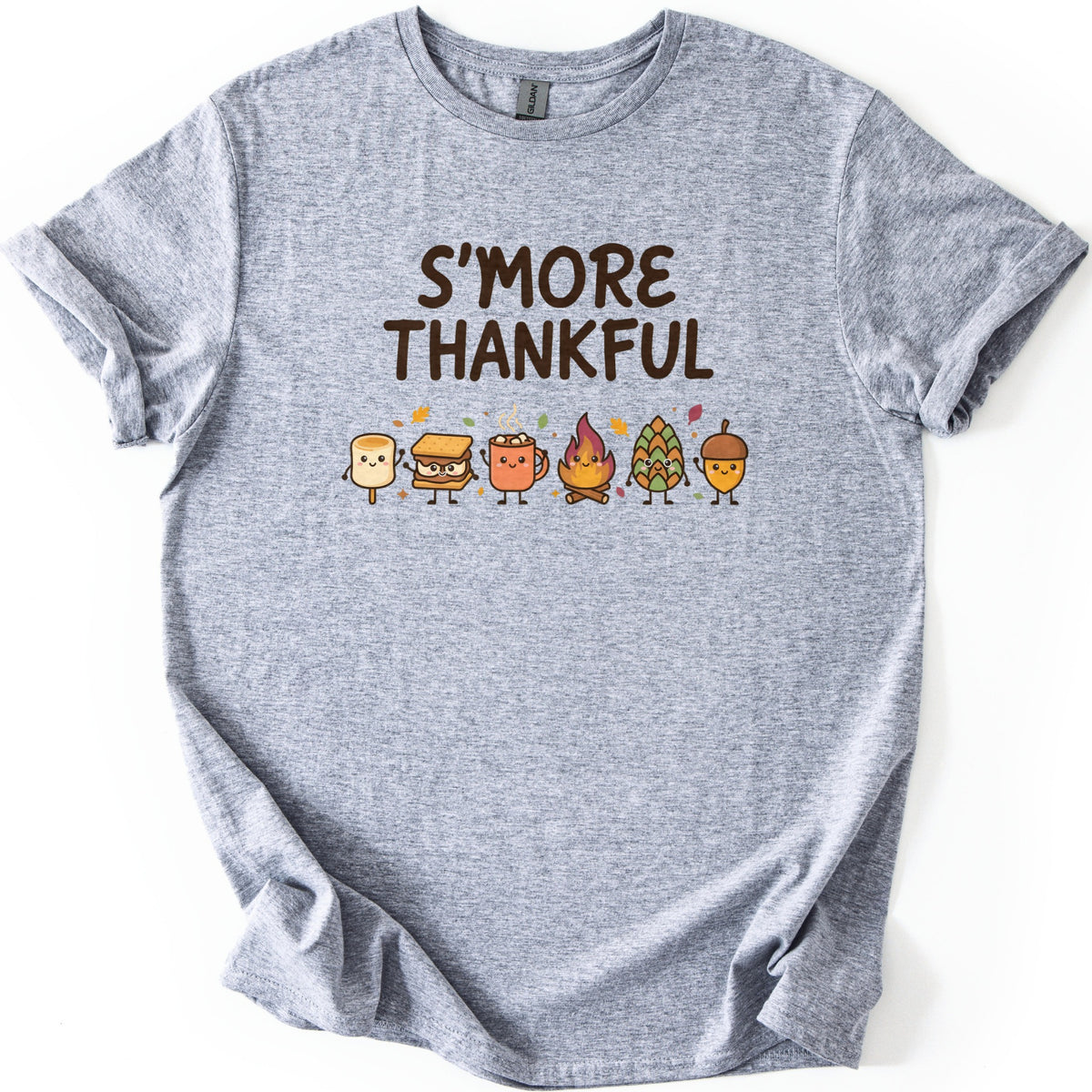 Smore Thankful Comfort Colors T-shirt in Sport Grey, laid flat. Dark brown letters read S'MORE THANKFUL above a row of smiling cartoon characters: a marshmallow, a s'more, a cocoa mug, a campfire, a pinecone and an acorn among autumn leaves. Sleeves rolled, on white.