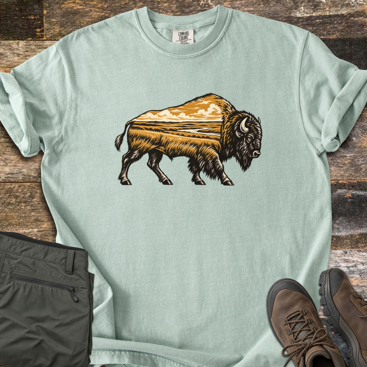 Prairie Bison Comfort Colors T-shirt in Bay, laid flat. A walking bison in black woodcut-style linework, its body filled with a golden prairie scene of grassland, a winding river and clouds. Folded olive cargo pants and brown hiking boots sit on weathered wood.