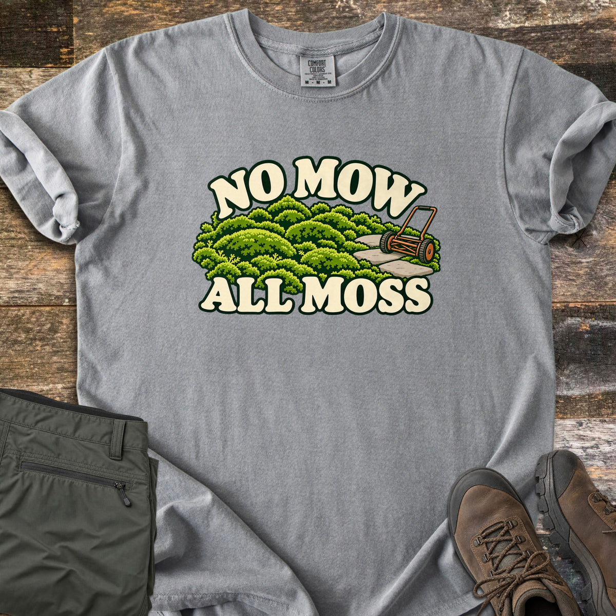 No Mow All Moss Comfort Colors T-shirt in Grey, laid flat. The print shows rounded green moss, stepping stones and an orange push reel mower framed by the cream words No Mow All Moss. It lies on rustic wood planks with folded olive pants and brown hiking boots.