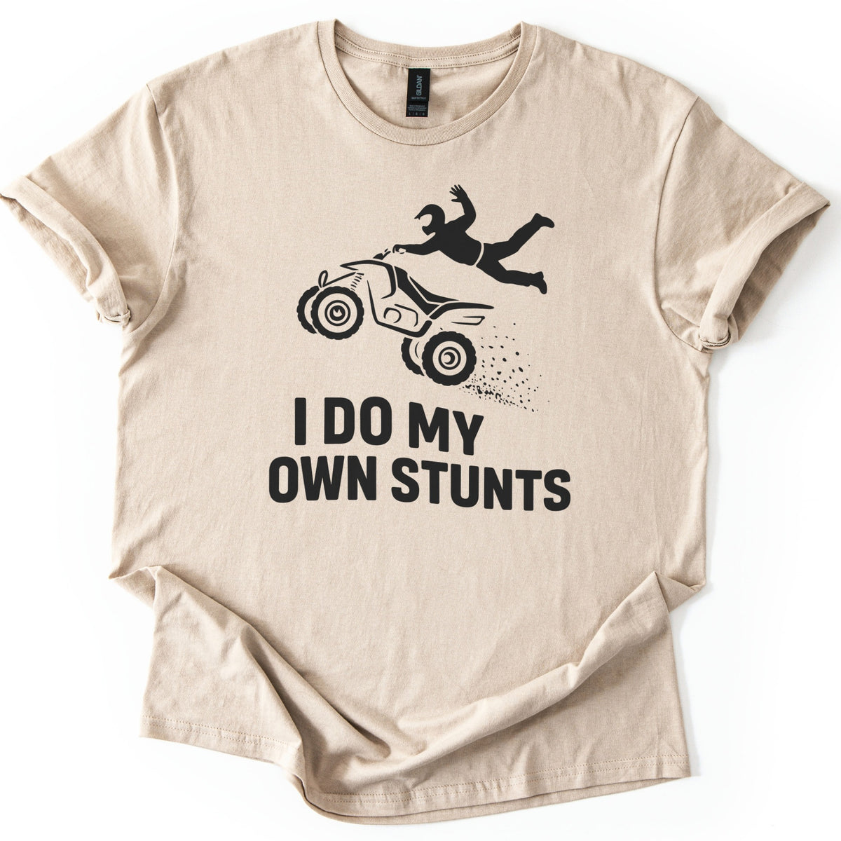 ATV Stunts Comfort Colors T-shirt in Sand, laid flat. A black silhouette of a helmeted rider flying off the back of an ATV as it kicks up dirt, above the words I DO MY OWN STUNTS. The shirt lies on a white background with its sleeves cuffed.