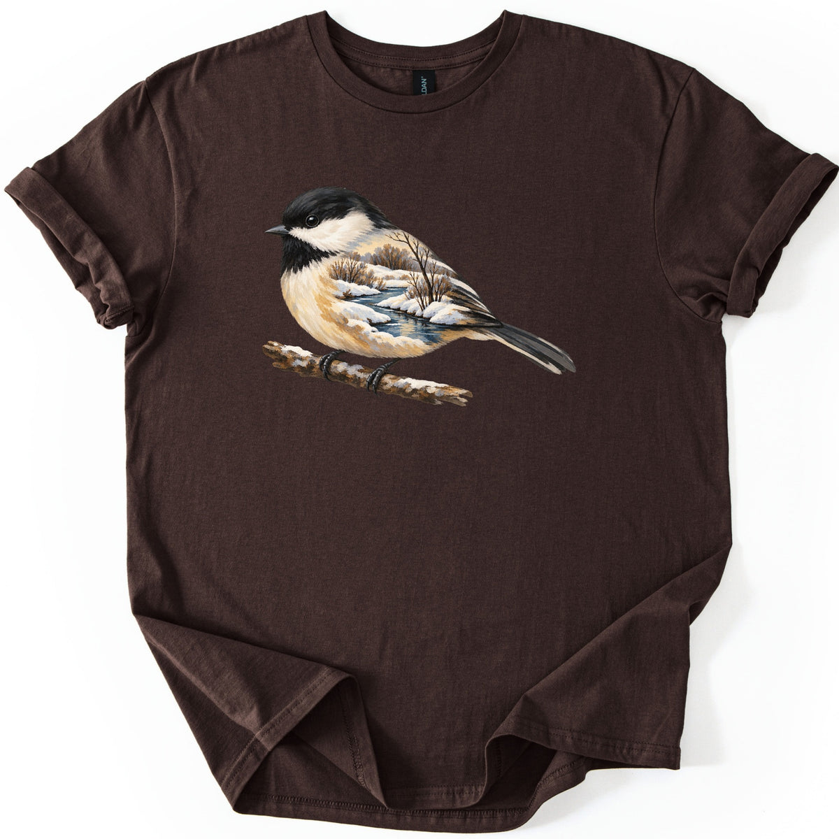 Chickadee Winter Creek Comfort Colors T-shirt in Dark Chocolate, laid flat. A painted black-capped chickadee perches on a snowy branch, its body filled with a winter scene of a creek, snowbanks and bare trees. No words are printed. The shirt lies on a white surface with rolled sleeves.