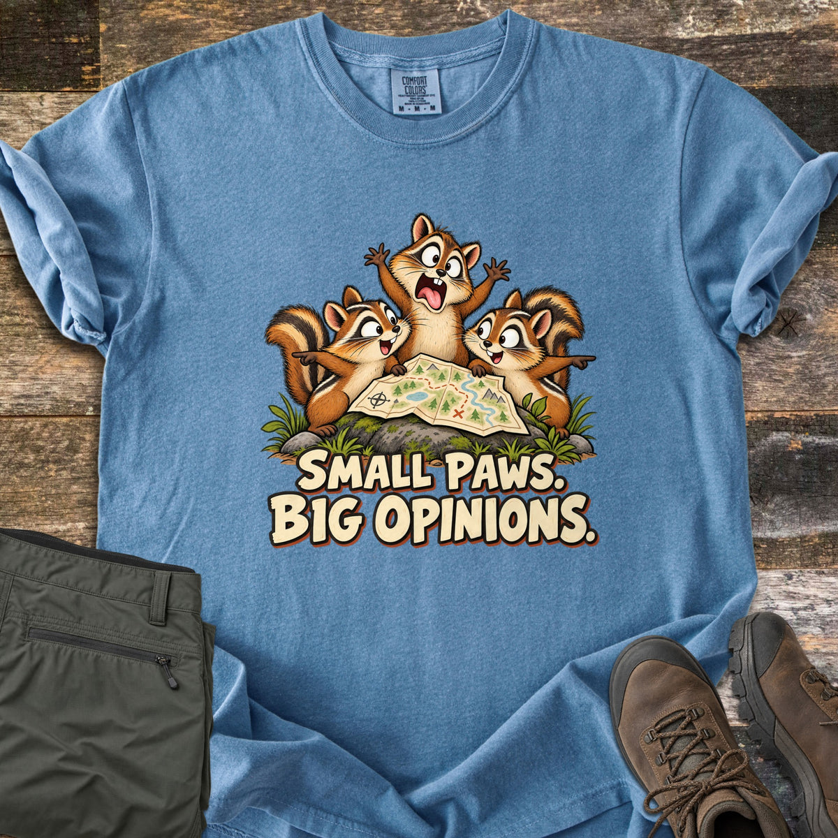 Small Paws. Big Opinions. Comfort Colors T-shirt in Blue Jean, laid flat. Three cartoon chipmunks gather around a trail map on a rock, two pointing in opposite directions, above the words SMALL PAWS. BIG OPINIONS. Folded olive pants and brown hiking boots rest on weathered boards.