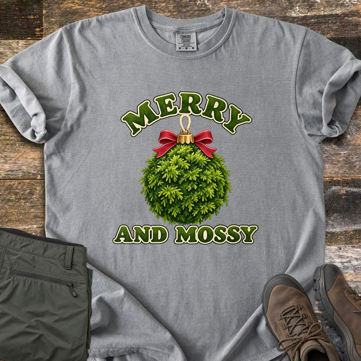Merry and Mossy Comfort Colors T-shirt in Grey, laid flat. A round Christmas ornament made of green moss, topped with a gold cap and red bow, sits between the words Merry and Mossy in green lettering. Olive hiking pants and brown hiking boots lie beside it on weathered wood.