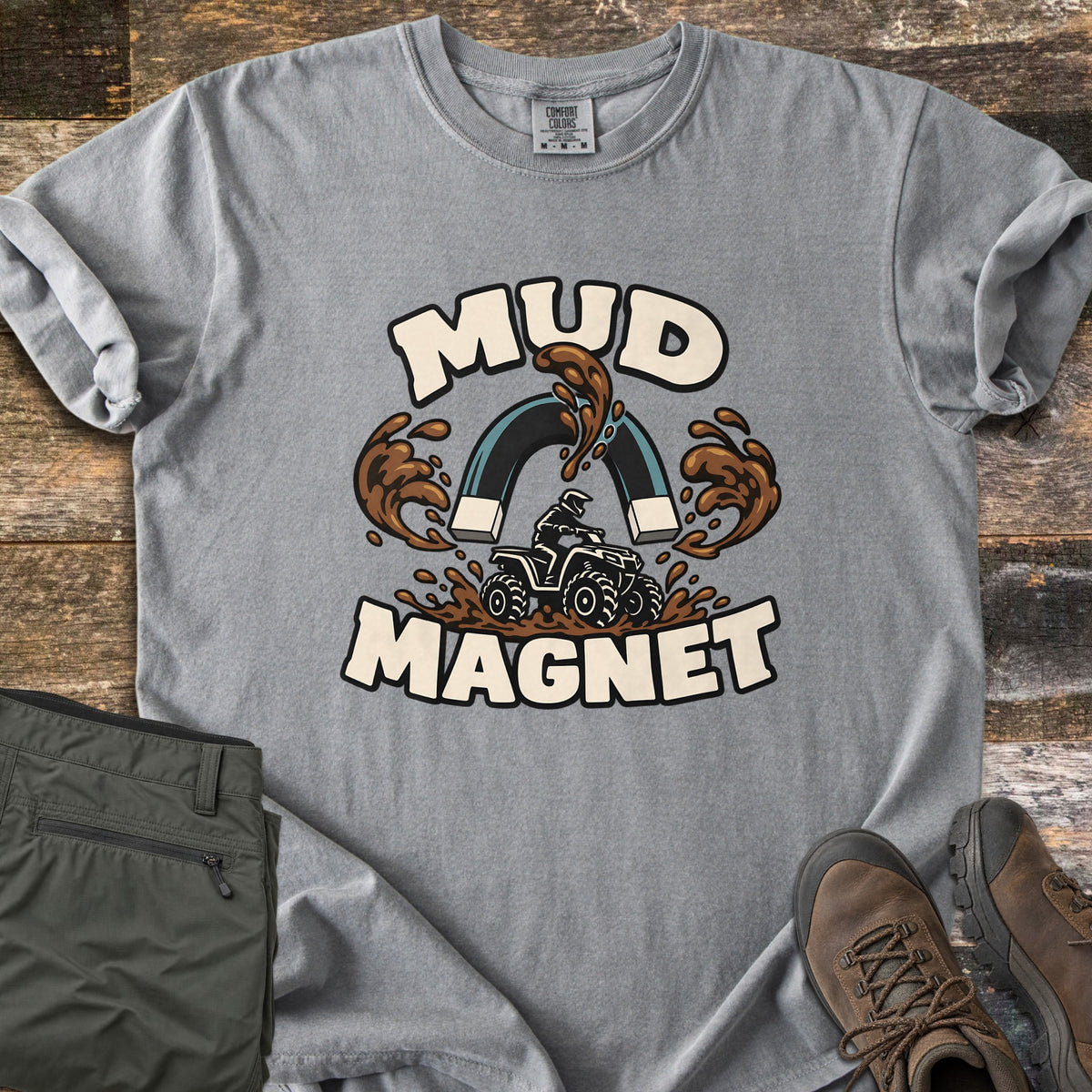 Mud Magnet Comfort Colors T-shirt in Grey, laid flat. The words MUD MAGNET arch around a horseshoe magnet pulling mud splashes toward an ATV rider churning through mud. Shown on a wood floor with folded olive pants and brown hiking boots.