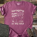 Foraging Fun-Guy in the Wild Comfort Colors T-shirt in Berry, laid flat. A cream line drawing of a capybara wearing a spotted mushroom cap, tongue out, sits in grass by tiny mushrooms between the words foraging FUN-GUY in the wild. Olive hiking pants and brown boots lie on weathered wood.