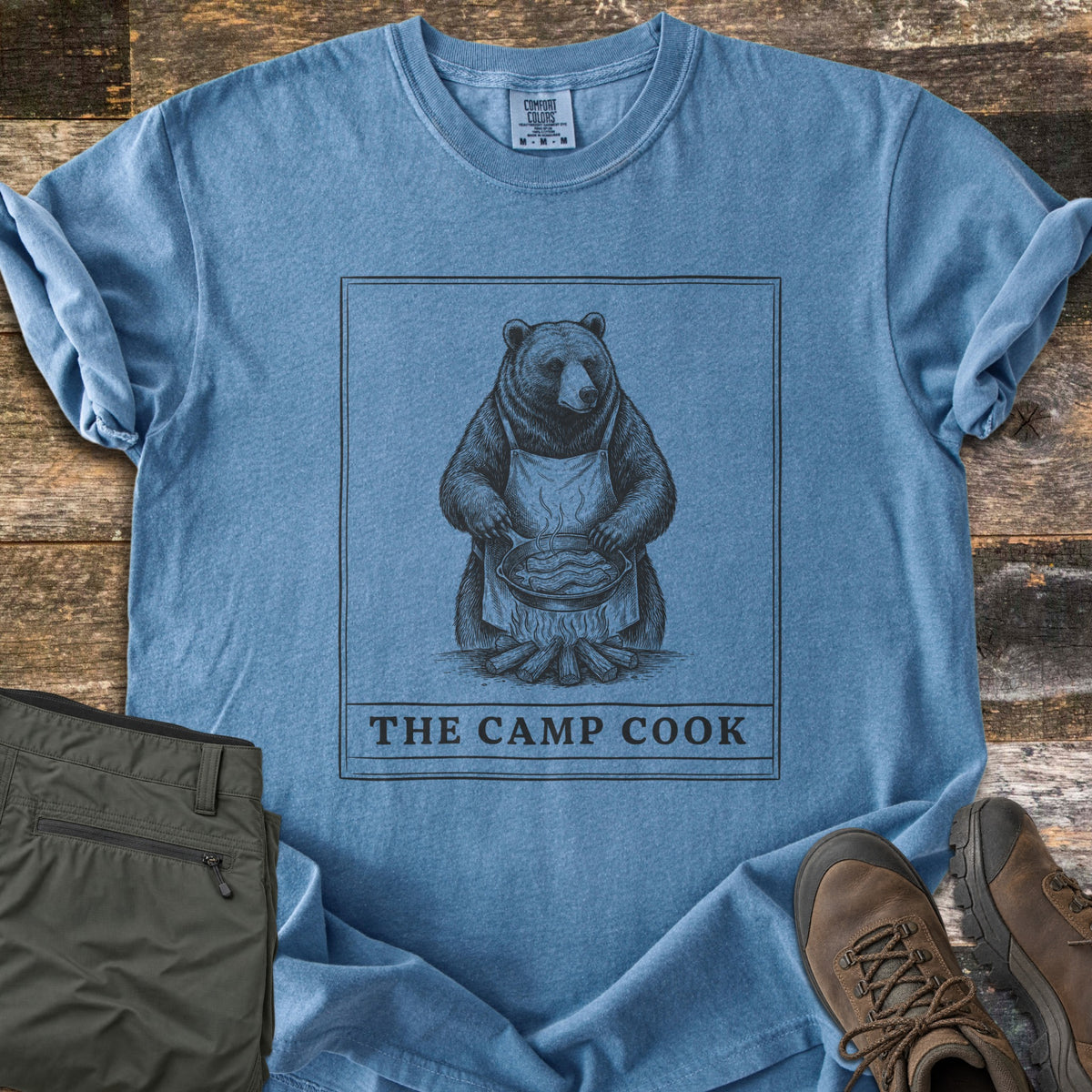 The Camp Cook Comfort Colors T-shirt in Blue Jean, laid flat. A framed sketch of a bear in an apron frying bacon in a skillet over a campfire, above the words THE CAMP COOK. Sleeves are cuffed on weathered wood beside olive hiking pants and brown hiking boots.