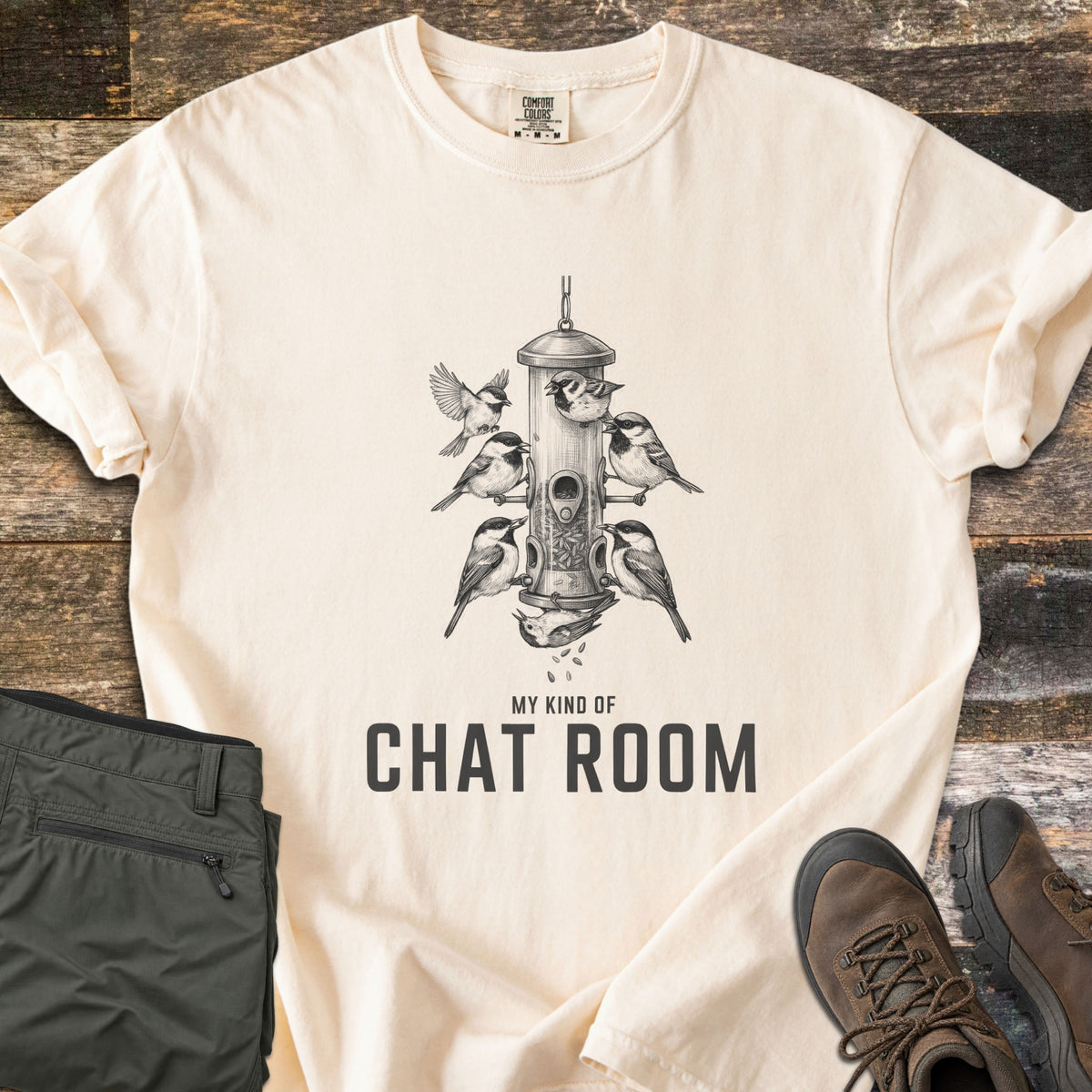 My Kind Of Chat Room Comfort Colors T-shirt in Ivory, laid flat. A dark gray sketch of chickadees perched around a hanging tube bird feeder, one upside down below it, above the words My Kind Of Chat Room. Olive hiking pants and brown boots lie beside it on weathered wood.