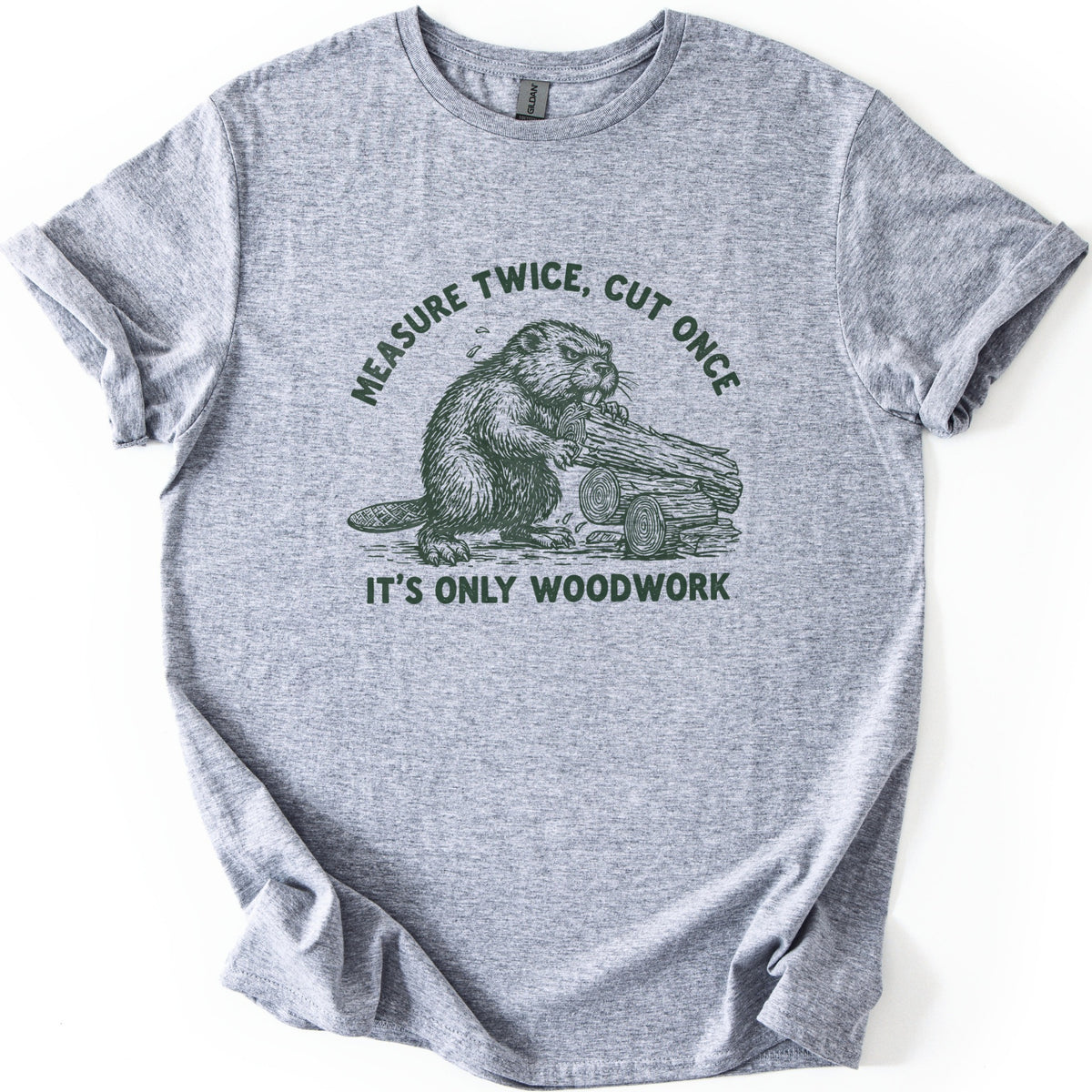 Measure Twice Beaver Comfort Colors T-shirt in Sport Grey, laid flat. A dark green sketch of a beaver gnawing a log beside cut logs, under the arched words Measure Twice, Cut Once and above It's Only Woodwork. The shirt has rolled sleeves on a white background.