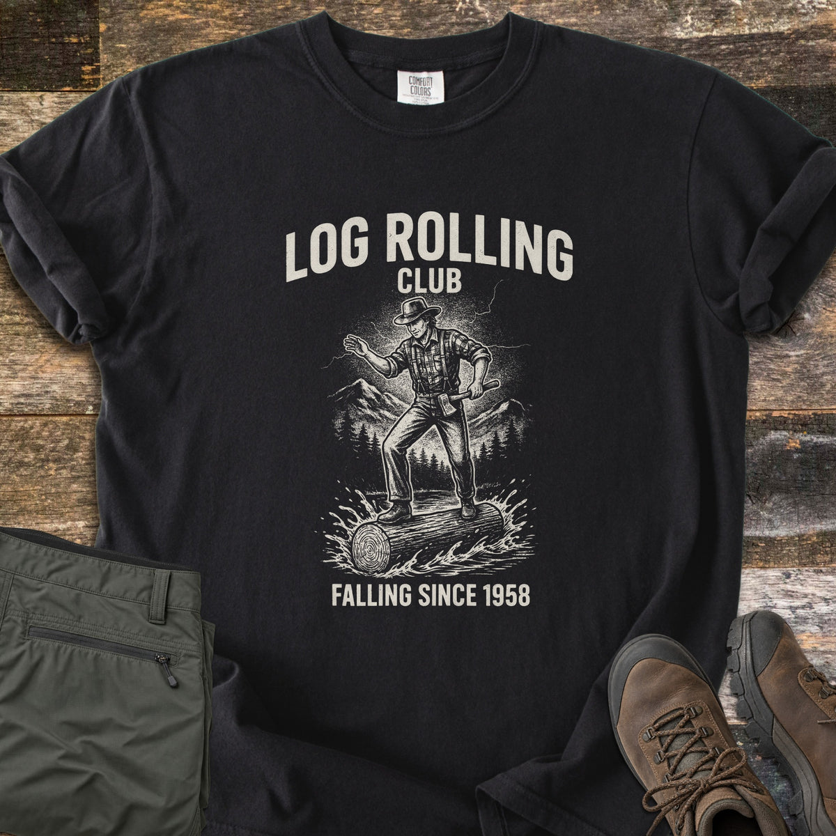 Log Rolling Club Comfort Colors T-shirt in Black, laid flat. A sketched lumberjack in a hat and plaid shirt, holding an axe, balances on a log splashing through water below the words Log Rolling Club and above Falling Since 1958. Olive pants and brown boots sit on wood planks.