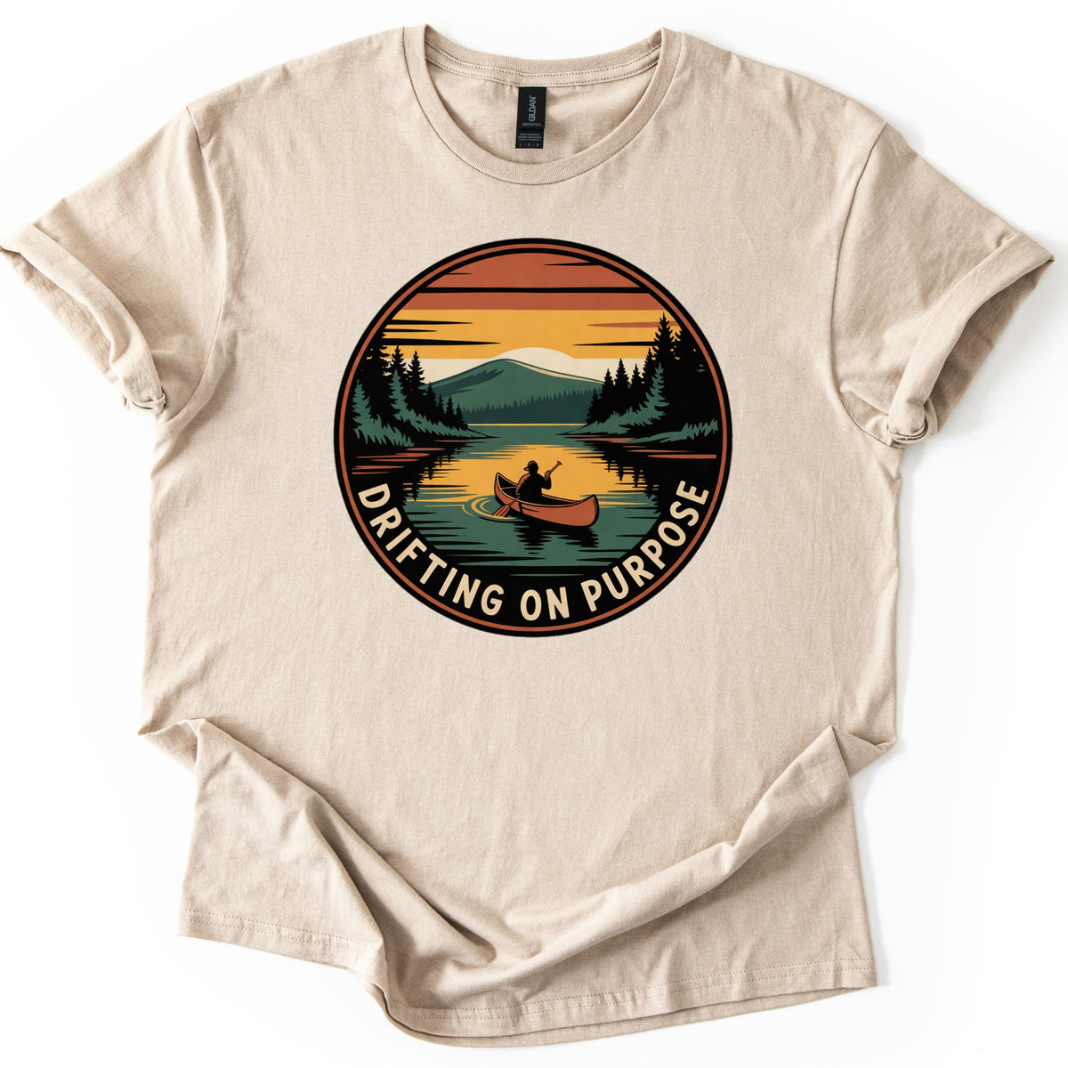 Drifting On Purpose Comfort Colors T-shirt in Sand, laid flat. A round badge shows a person paddling a red canoe on a lake at sunset, with pine trees and a green mountain, above the curved words Drifting On Purpose. The sleeves are cuffed on a white background.