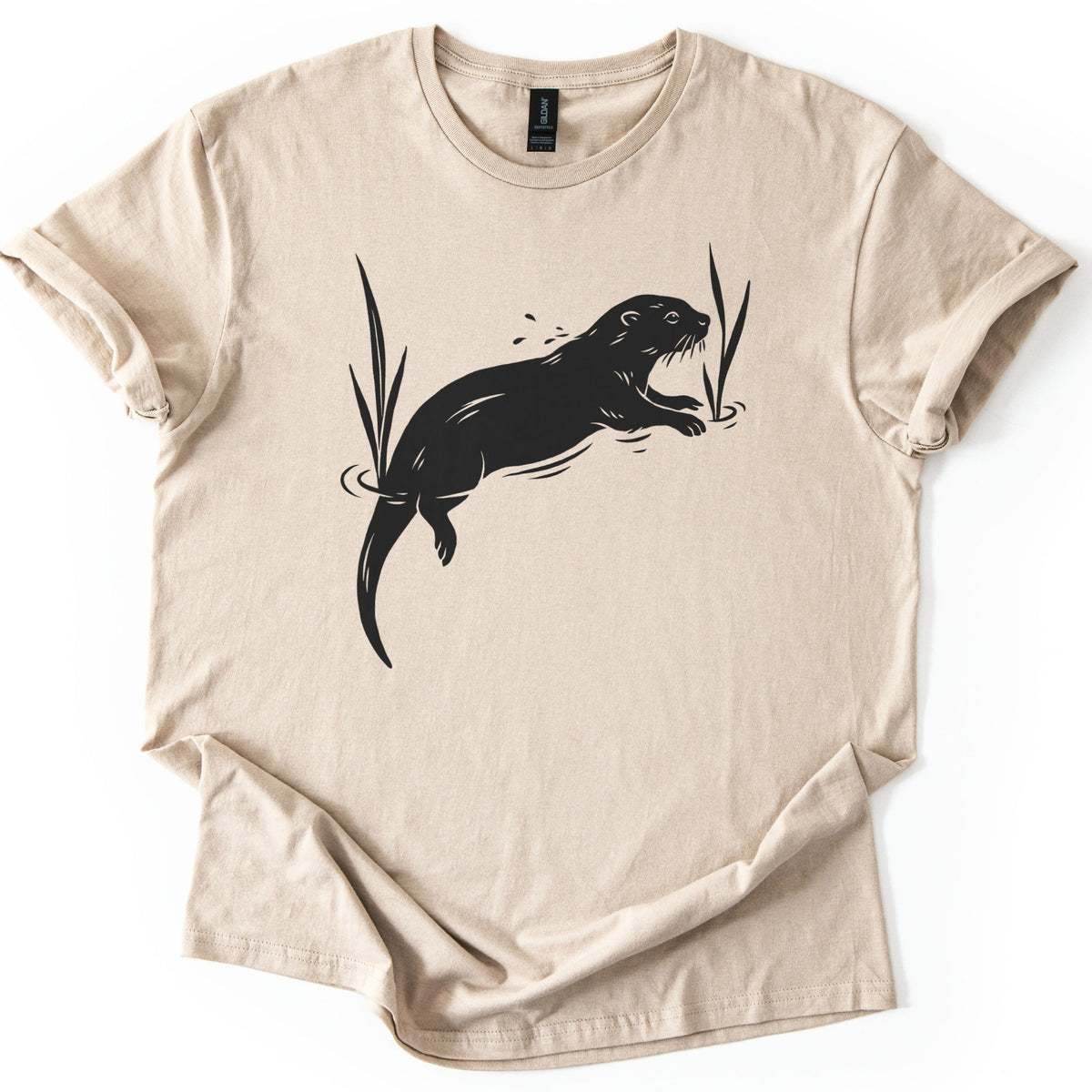 Swimming Otter Comfort Colors T-shirt in Sand, laid flat. A black linocut-style otter swims through rippling water between tall reeds, with droplets above its head. No words are printed. The sleeves are cuffed and the shirt lies on a white background.