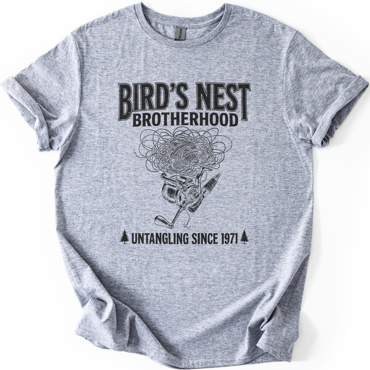 Birds Nest Brotherhood Comfort Colors T-shirt in Sport Grey, laid flat. Black print reads Bird's Nest Brotherhood above a tangled mess of fishing line over a spinning reel, rod and hook, then Untangling Since 1971 between two pine trees. Sleeves cuffed, on a white backdrop.
