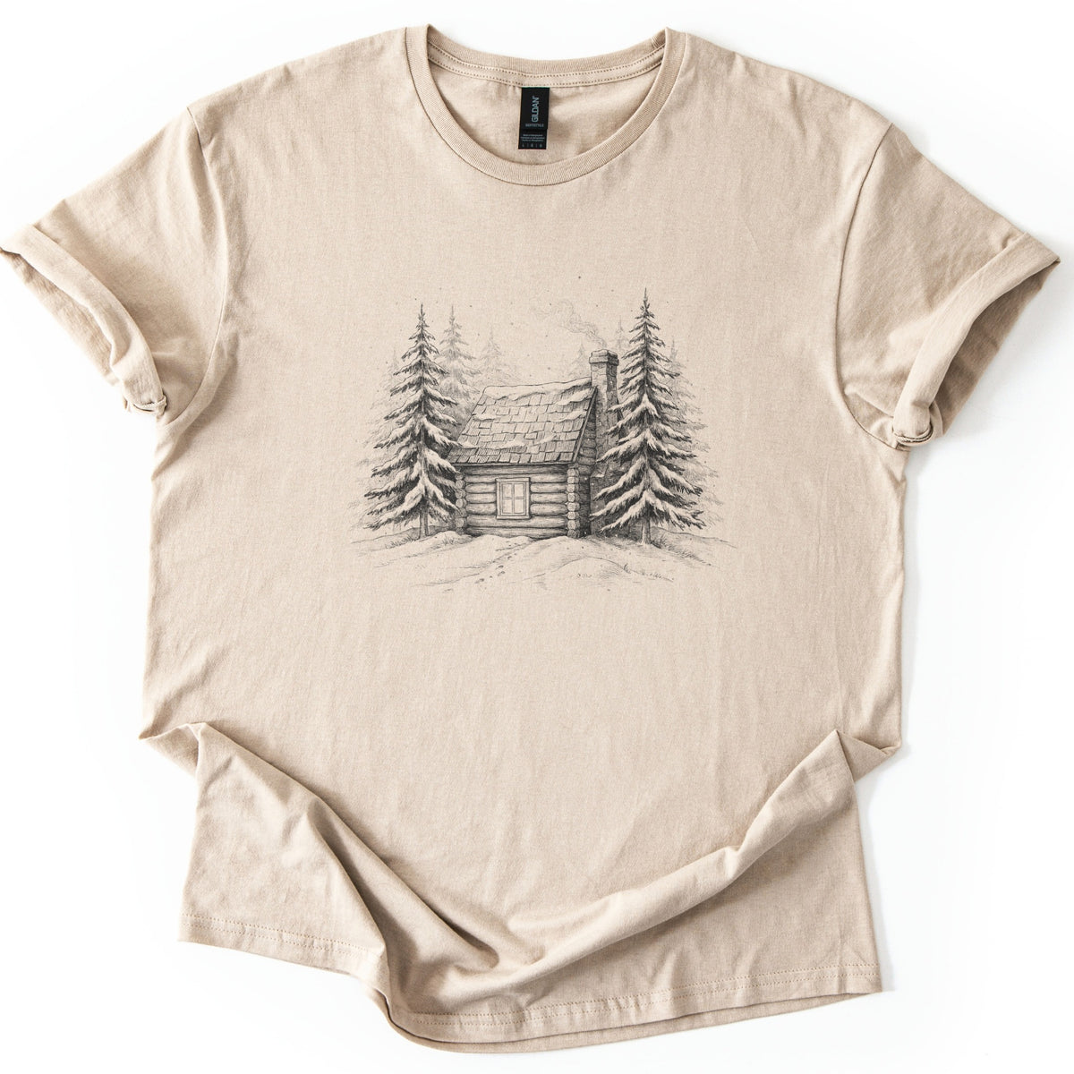 Snowy Log Cabin Comfort Colors T-shirt in Sand, laid flat. A pencil-style sketch of a snow-covered log cabin with smoke rising from its chimney, set between snowy pine trees on a drift. No words. Sleeves are rolled on a white background.