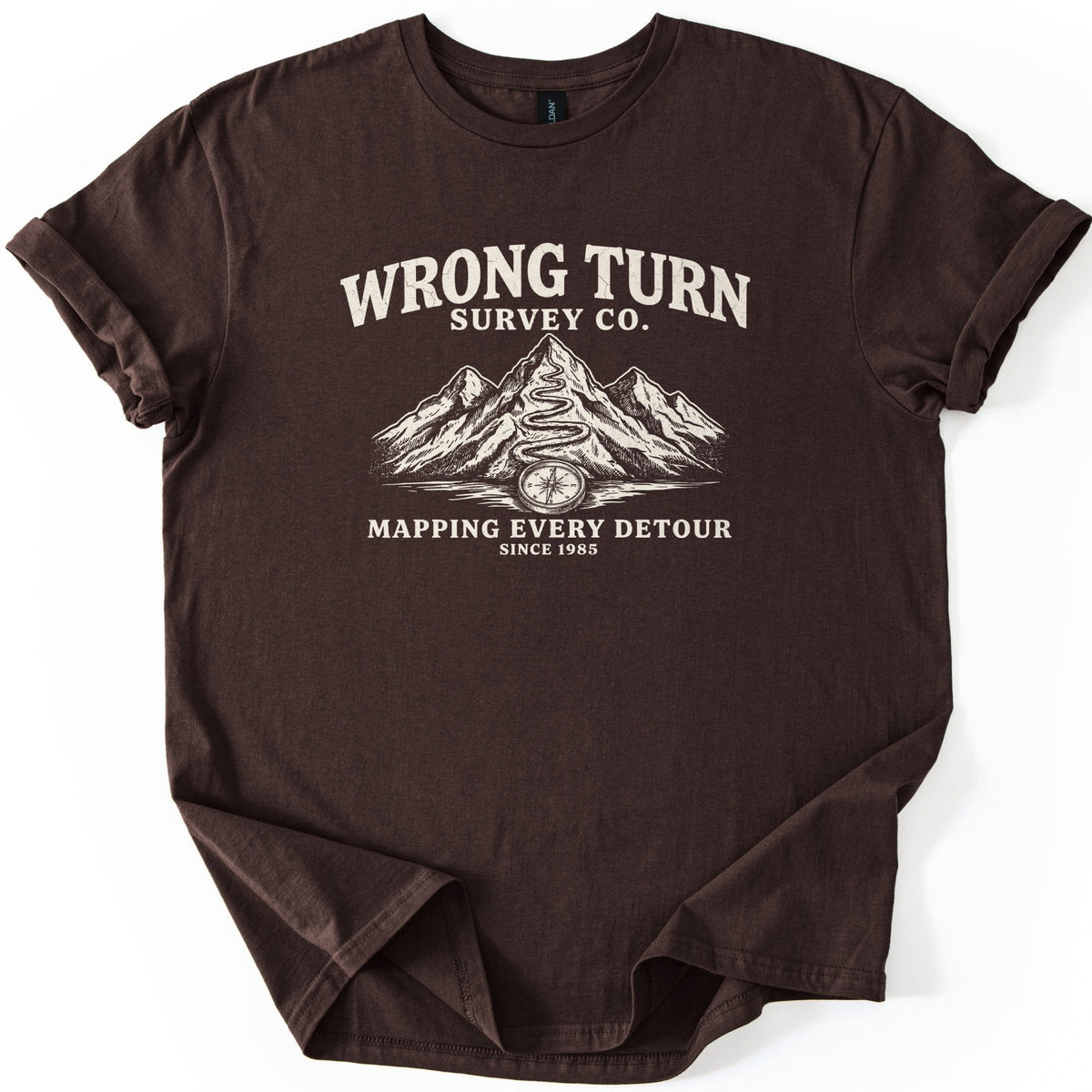 Wrong Turn Survey Co Comfort Colors T-shirt in Dark Chocolate, laid flat. Cream print reads Wrong Turn Survey Co. above sketched mountains with a winding trail up the tallest peak and a compass at its base, then Mapping Every Detour, Since 1985. Sleeves cuffed on a white background.