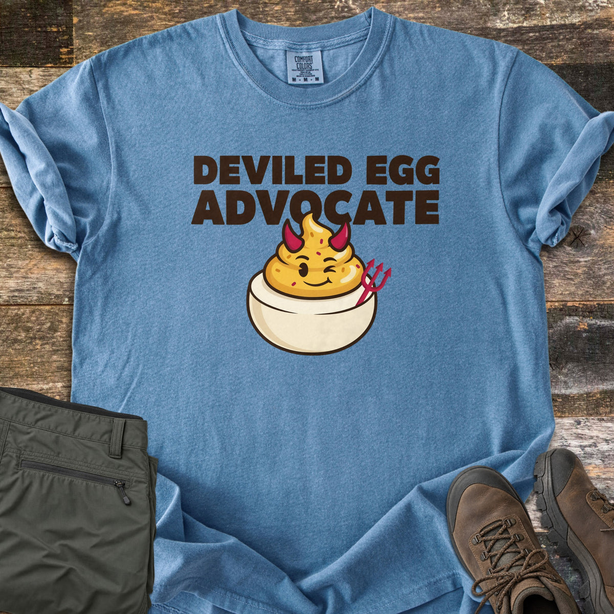 Deviled Egg Advocate Comfort Colors T-shirt in Blue Jean, laid flat. Dark brown letters read Deviled Egg Advocate above a winking cartoon deviled egg with red devil horns and a red pitchfork. Folded olive hiking pants and brown hiking boots sit on weathered wood planks.