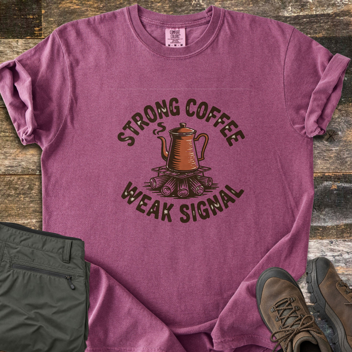Strong Coffee Weak Signal Comfort Colors T-shirt in Berry, laid flat. A steaming copper coffee pot sits on a grate over campfire logs, arched by the words Strong Coffee above and Weak Signal below. Folded olive hiking pants and brown hiking boots lie on weathered wood planks.