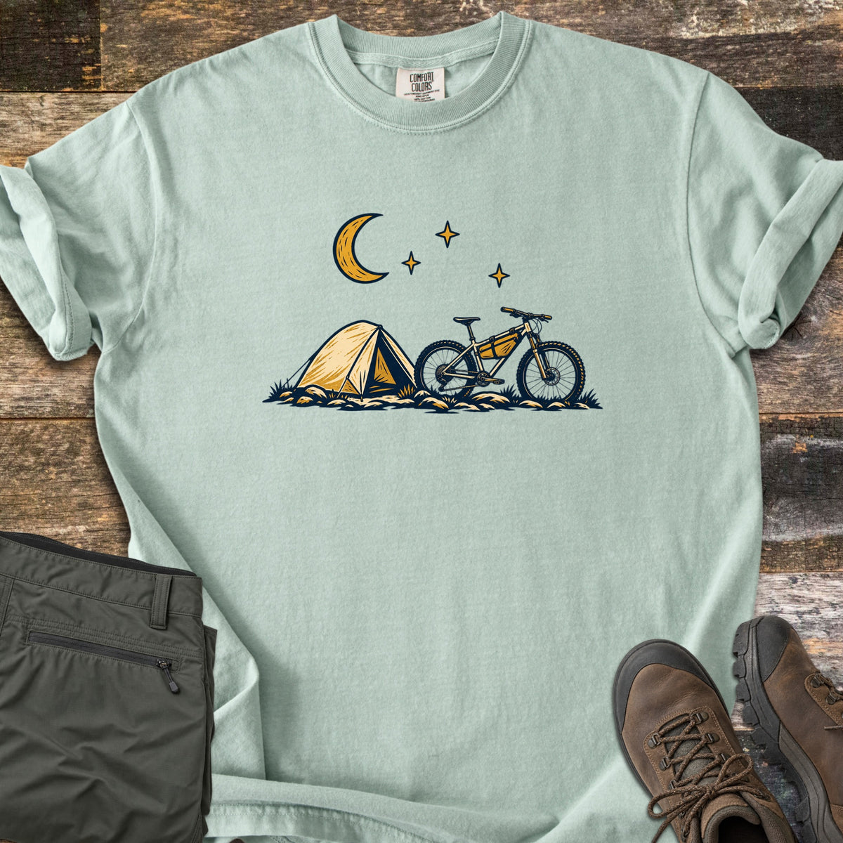 Bikepacking Under the Stars Comfort Colors T-shirt in Bay, laid flat. A yellow tent and a mountain bike with a frame bag sit on grassy ground under a crescent moon and three stars, with no words. Olive hiking pants and brown boots lie beside it on weathered wood planks.
