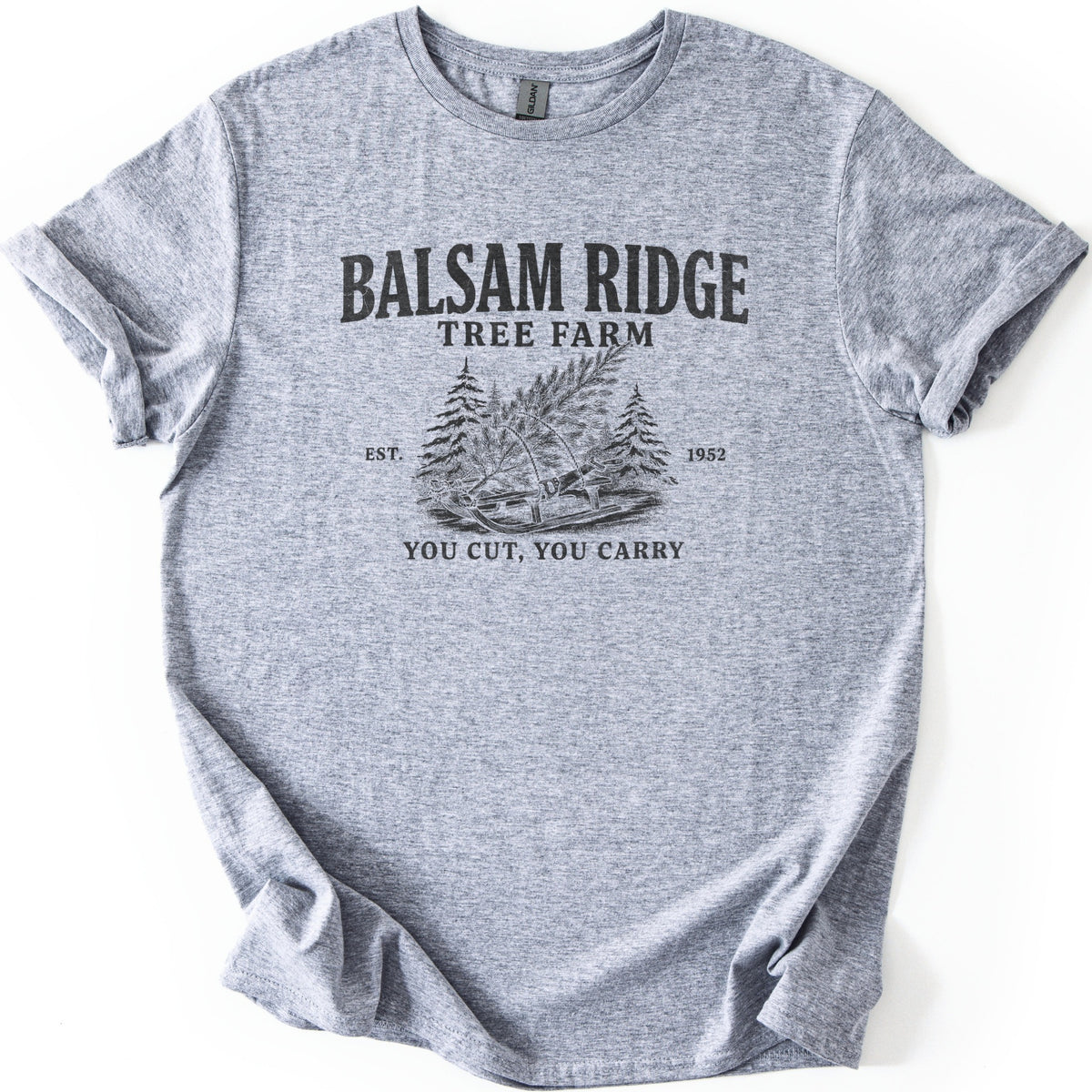 Balsam Ridge Tree Farm Comfort Colors T-shirt in Sport Grey, laid flat. Black print reads Balsam Ridge Tree Farm above an engraved-style fir tree tied to a wooden sled between pines, flanked by Est. 1952, with You Cut, You Carry below. Sleeves cuffed on a white surface.
