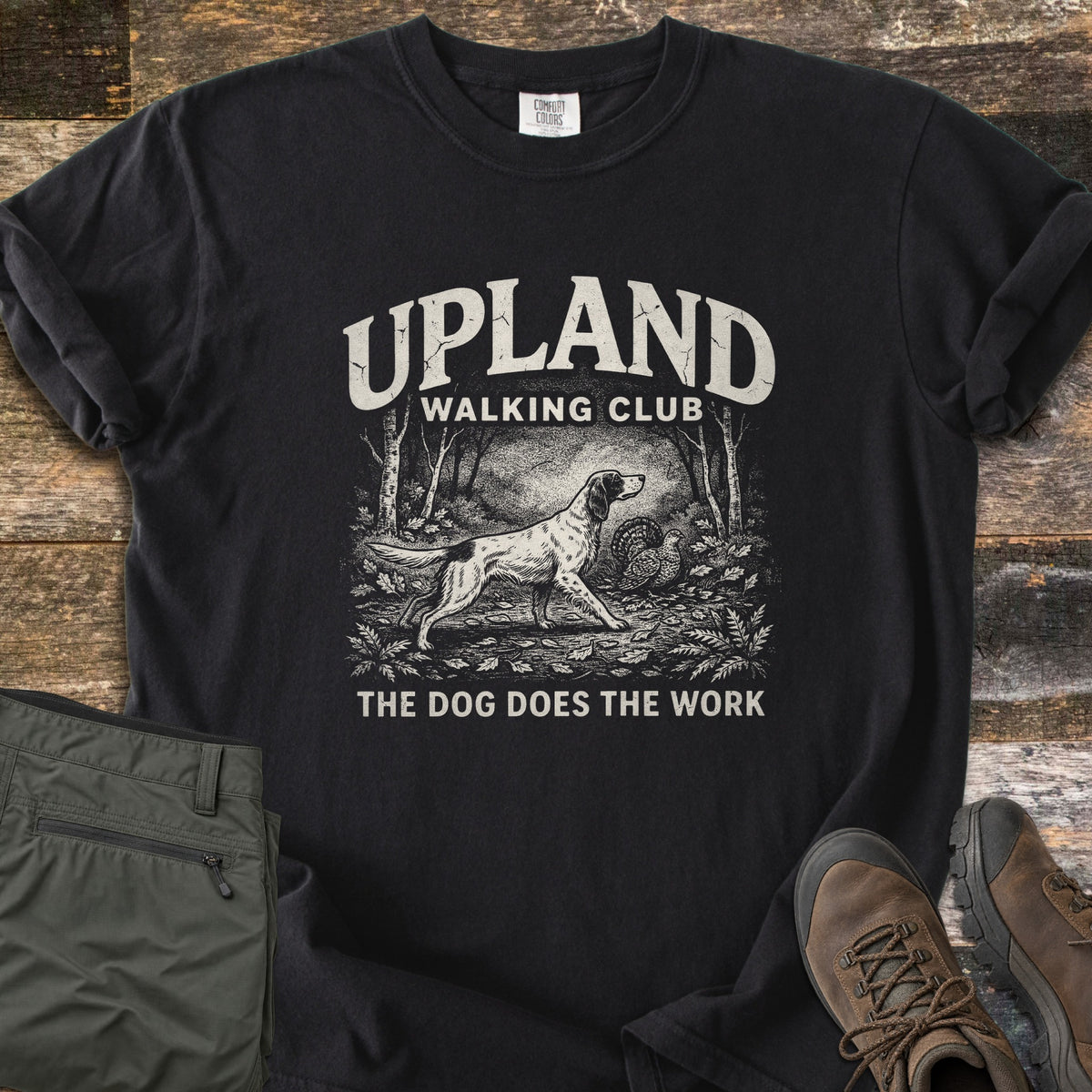 Upland Walking Club Comfort Colors T-shirt in Black, laid flat. A vintage-style sketch of a spaniel pointing at a grouse in the woods, under the words Upland Walking Club and above The Dog Does The Work. Olive pants and brown hiking boots sit beside it on weathered wood.
