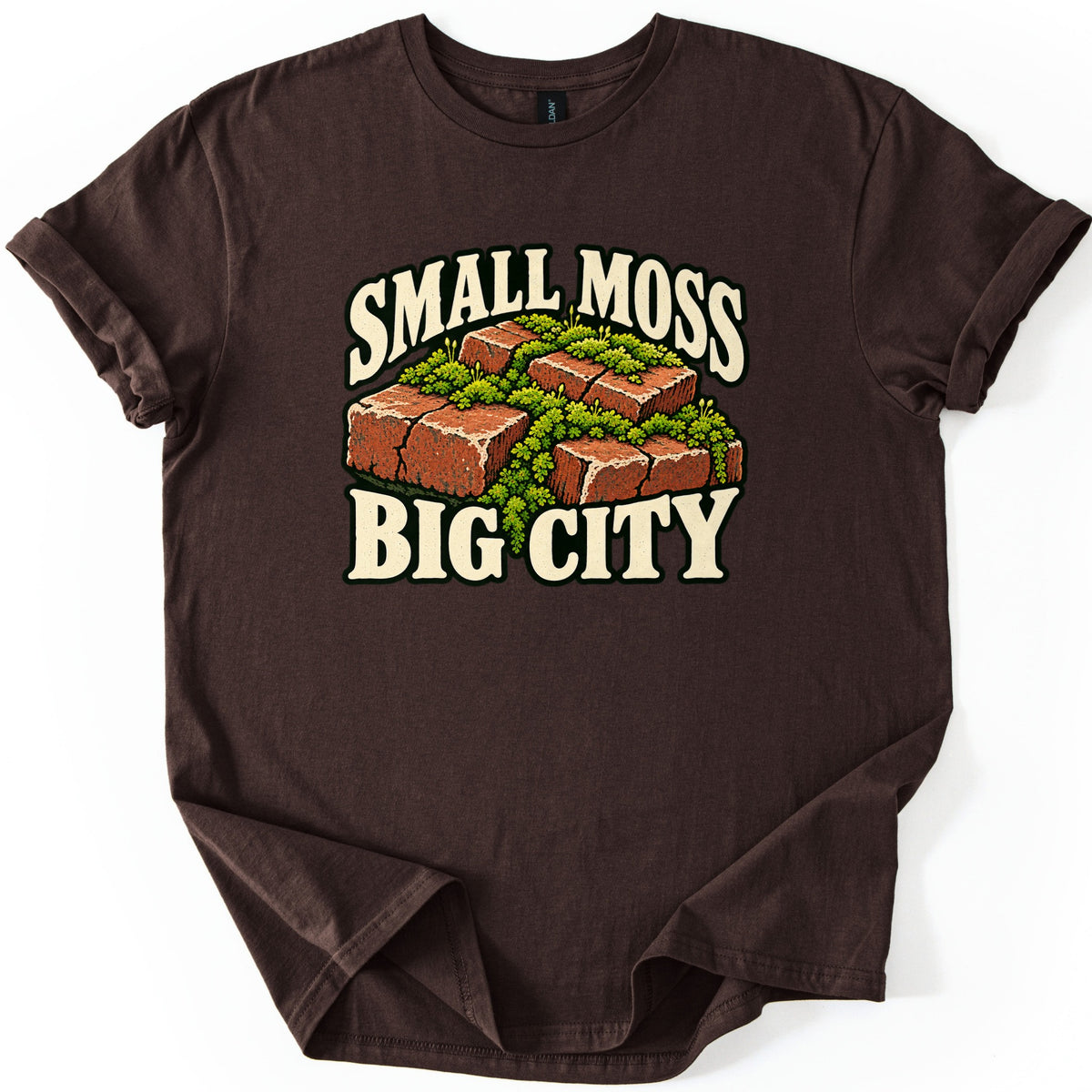 Small Moss Big City Comfort Colors T-shirt in Dark Chocolate, laid flat. A cluster of red bricks overgrown with green moss sits between the words SMALL MOSS and BIG CITY in cream letters. The shirt has rolled sleeves and lies on a white background.