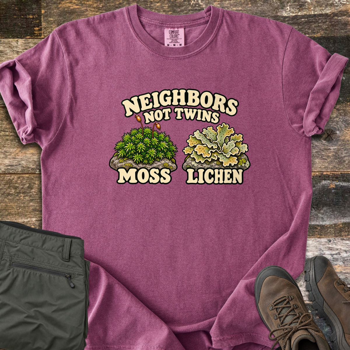 Neighbors Not Twins Comfort Colors T-shirt in Berry, laid flat. Arched text Neighbors Not Twins over two illustrated rock clumps: green moss with brown stalks labeled Moss, and pale green leafy lichen labeled Lichen. Olive hiking pants and brown boots sit beside it on weathered wood.