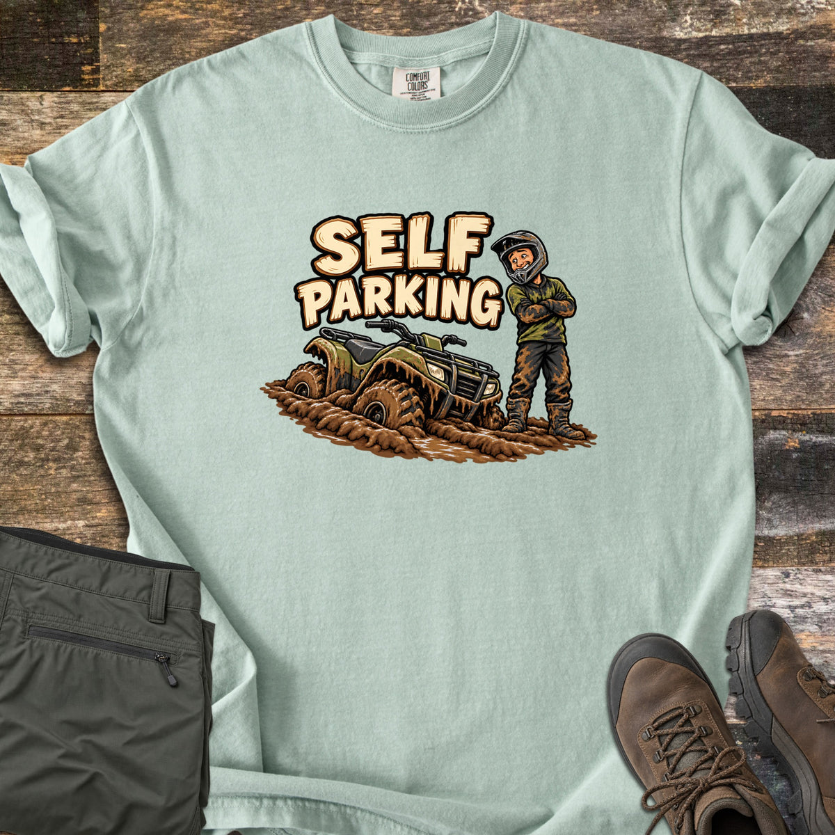 Self Parking Comfort Colors T-shirt in Bay, laid flat. A cartoon rider in a helmet stands arms crossed, covered in mud, beside an ATV sunk in mud under the words Self Parking. Folded olive pants and brown hiking boots sit on a wood plank background.