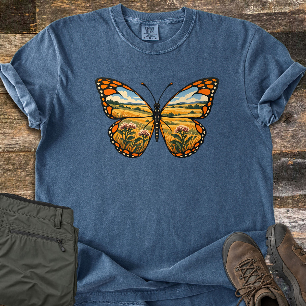 Monarch Prairie Comfort Colors T-shirt in Denim, laid flat. A monarch butterfly whose wings show a golden prairie scene with blue sky, clouds and pink milkweed blooms. The shirt lies on weathered wood beside olive green pants and brown hiking boots.