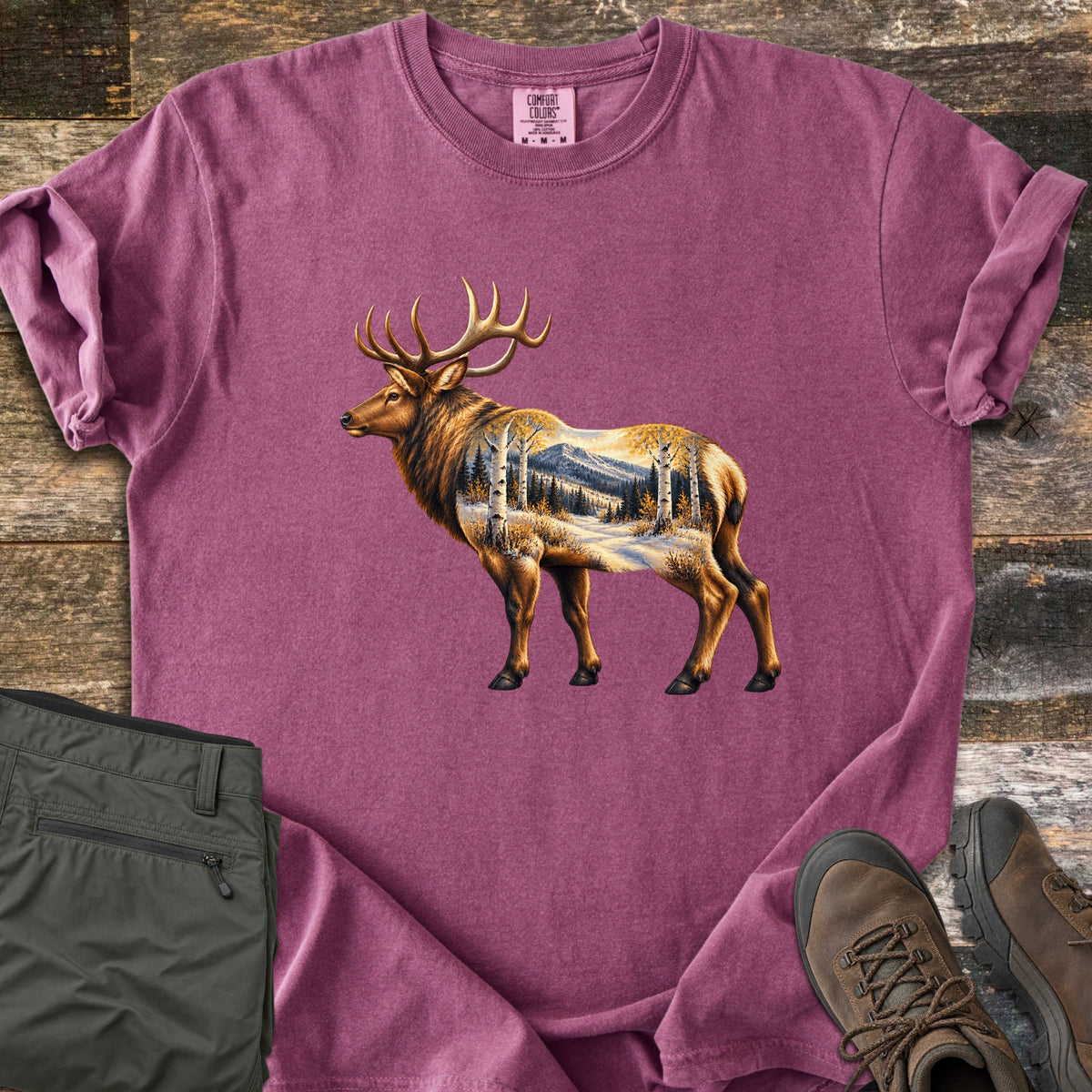 Aspen Pass Elk Comfort Colors T-shirt in Berry, laid flat. A painted bull elk with large antlers, its body filled with a winter scene of aspens, pines and a snowy mountain. Sleeves cuffed, on weathered wood with olive hiking pants and brown hiking boots.