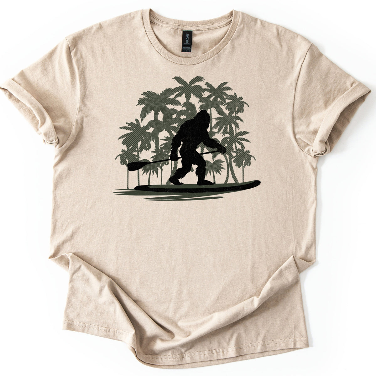 Paddle Board Squatch Comfort Colors T-shirt in Sand, laid flat. A black silhouette of Bigfoot stands on a paddle board holding a paddle, in front of a cluster of olive green palm trees, with no words printed. The shirt lies on a white background with rolled sleeves.