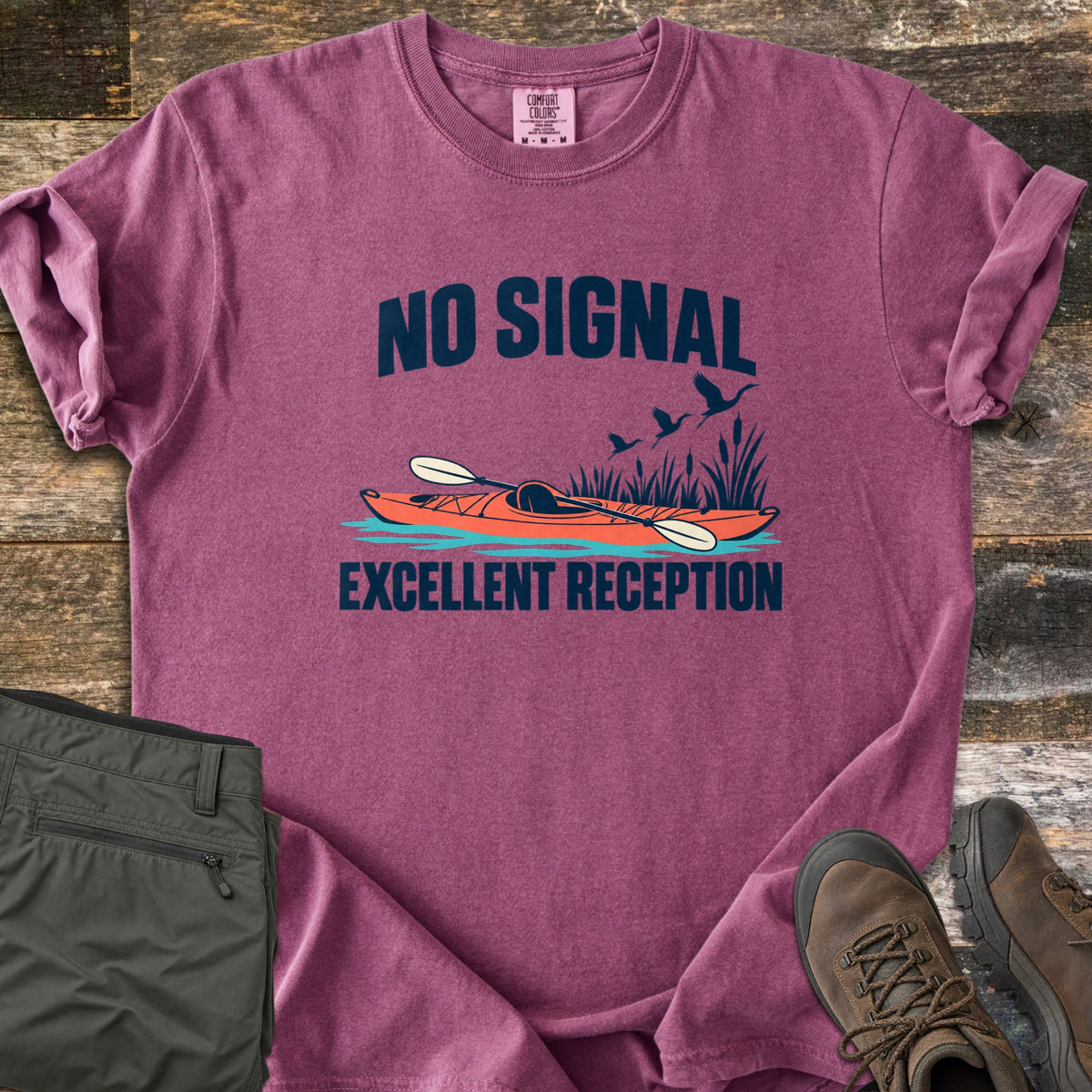 Excellent Reception Comfort Colors T-shirt in Berry, laid flat. The words No Signal arch over an orange kayak with a paddle among reeds and flying birds, above the words Excellent Reception. Olive pants and brown hiking boots sit on a weathered wood surface.