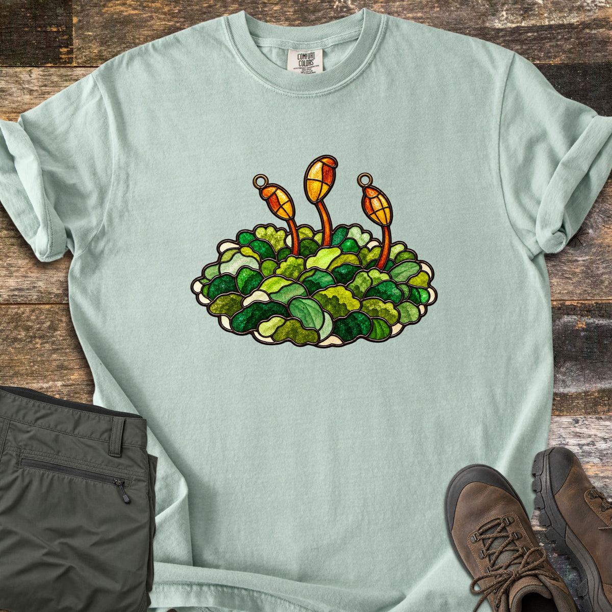 Stained Glass Moss Comfort Colors T-shirt in Bay, laid flat. Green moss in stained glass style with three orange and yellow spore capsules on red stalks, no words. The sleeves are rolled, with folded olive pants and brown hiking boots on dark wood boards.