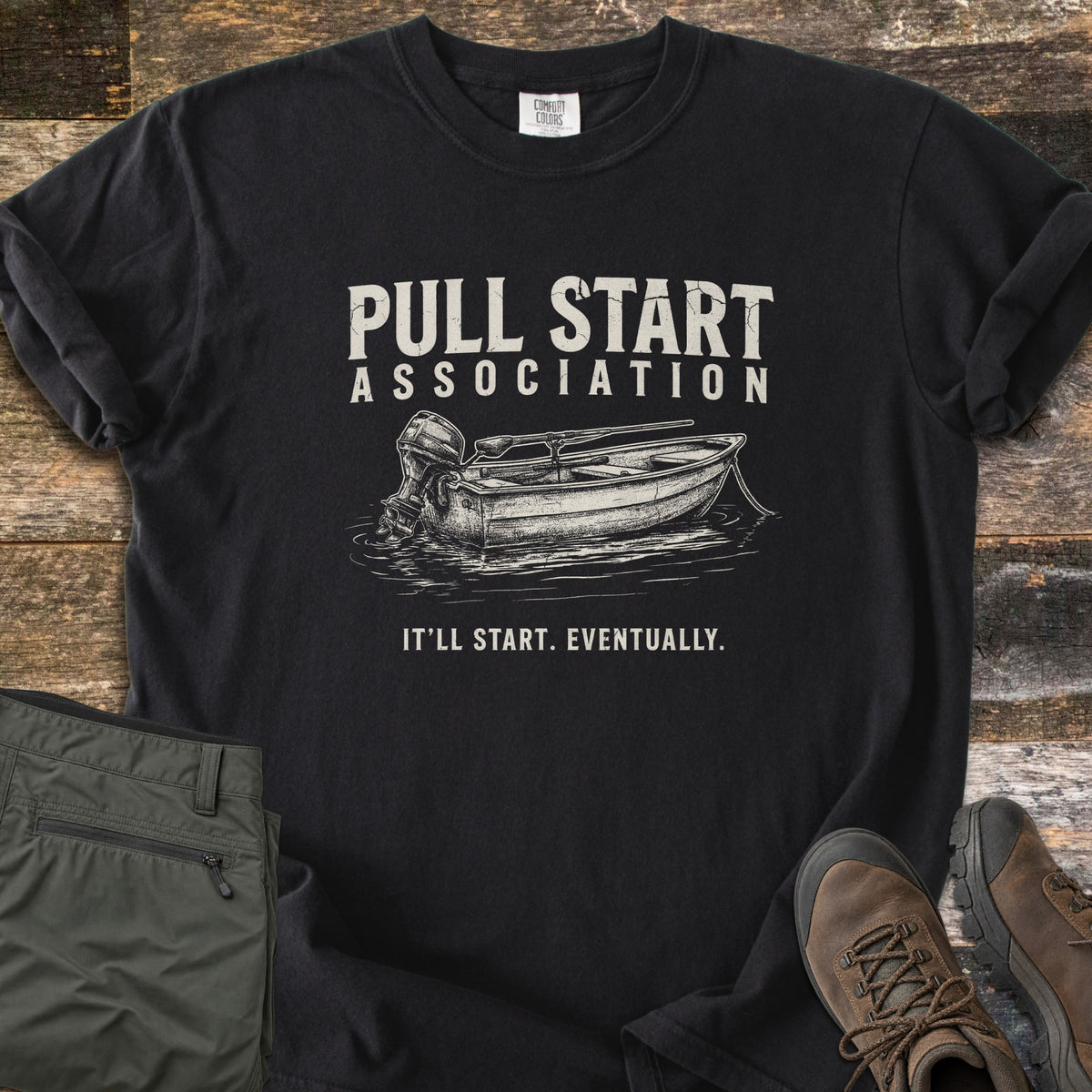 Pull Start Association Comfort Colors T-shirt in Black, laid flat. Cream distressed text reads Pull Start Association above a sketched rowboat with an outboard motor on water, and below it It'll Start. Eventually. Sleeves are rolled on a wood floor beside green pants and brown hiking boots.