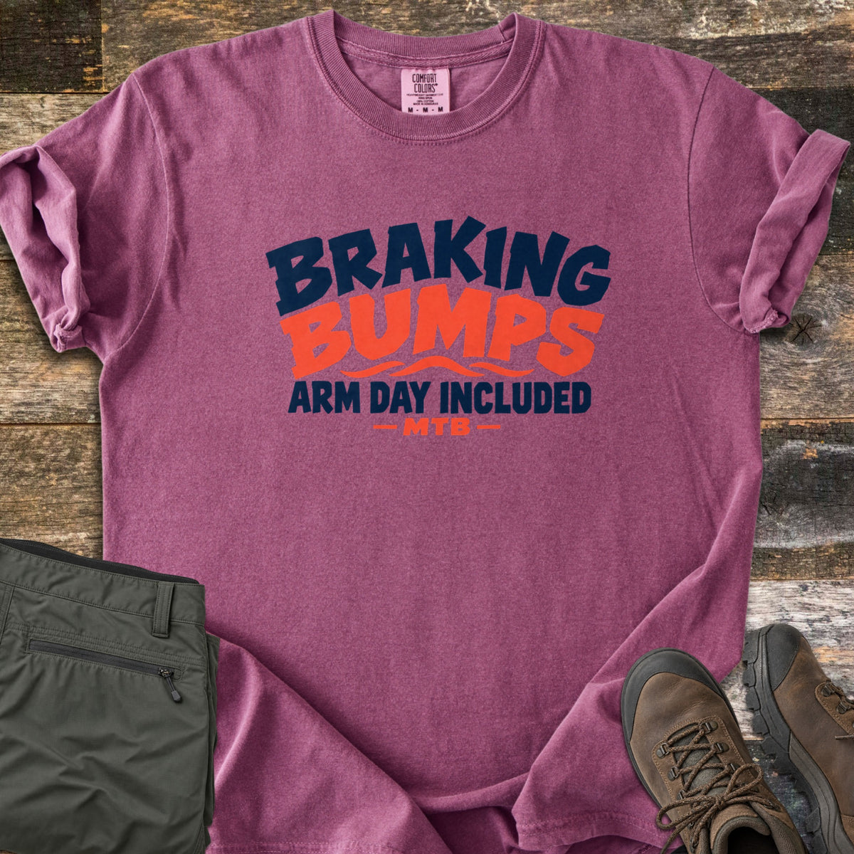 Braking Bumps Arm Day Included Comfort Colors T-shirt in Berry, laid flat. Bold navy and orange text reads Braking Bumps, Arm Day Included, MTB, with wavy orange lines under Bumps. It rests on a wood floor beside olive cargo pants and brown hiking boots.