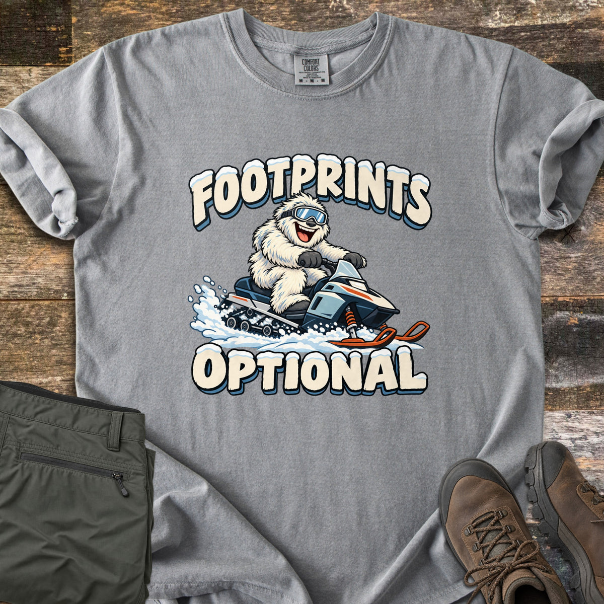 Footprints Optional Comfort Colors T-shirt in Grey, laid flat. A grinning white yeti in ski goggles rides a snowmobile through spraying snow between the snow-capped words Footprints Optional. It rests on weathered wood planks next to folded olive pants and brown hiking boots.