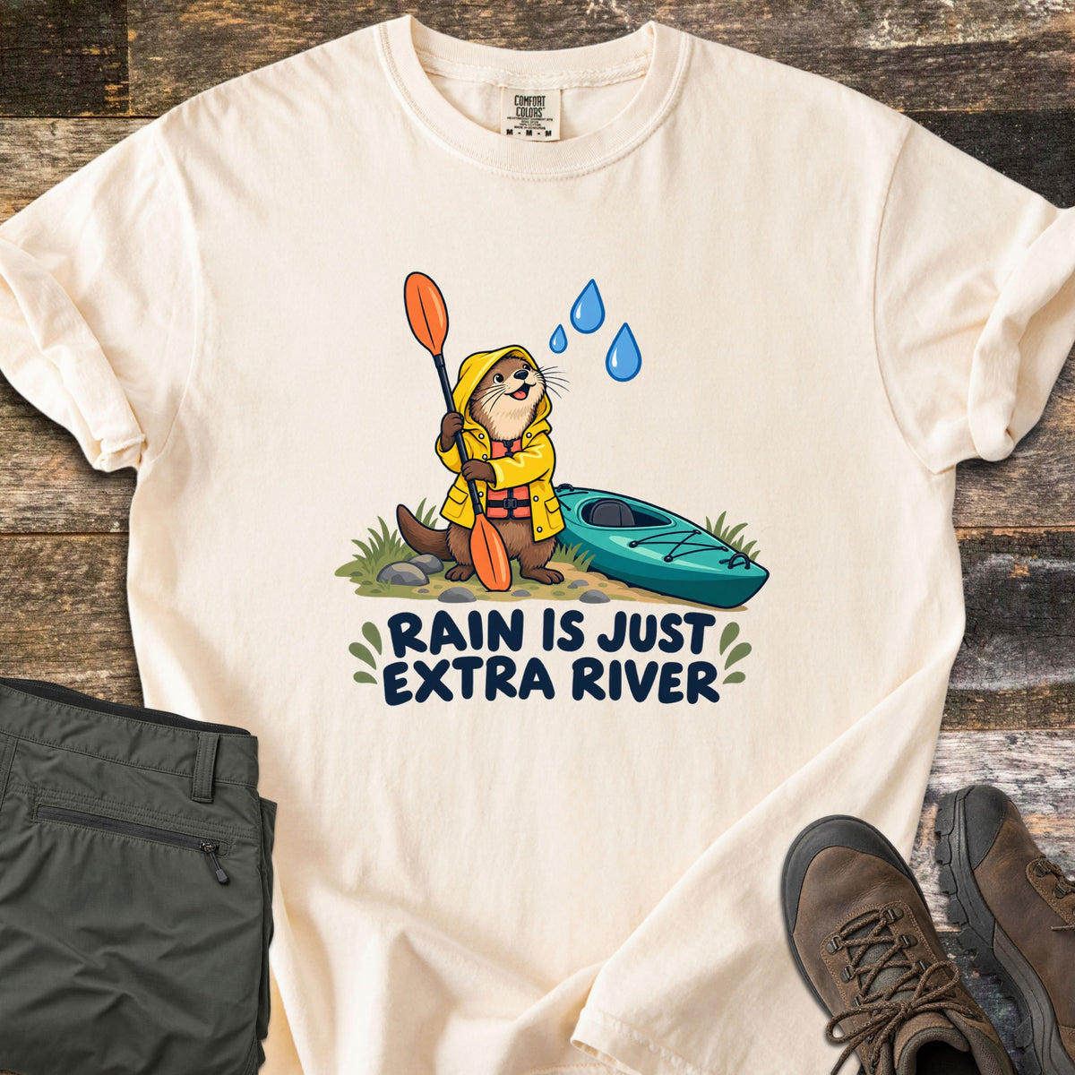 Extra River Comfort Colors T-shirt in Ivory, laid flat. A cartoon otter in a yellow raincoat and life vest holds an orange paddle beside a teal kayak as raindrops fall, above the words Rain Is Just Extra River. Olive pants and brown hiking boots lie on a wood floor.