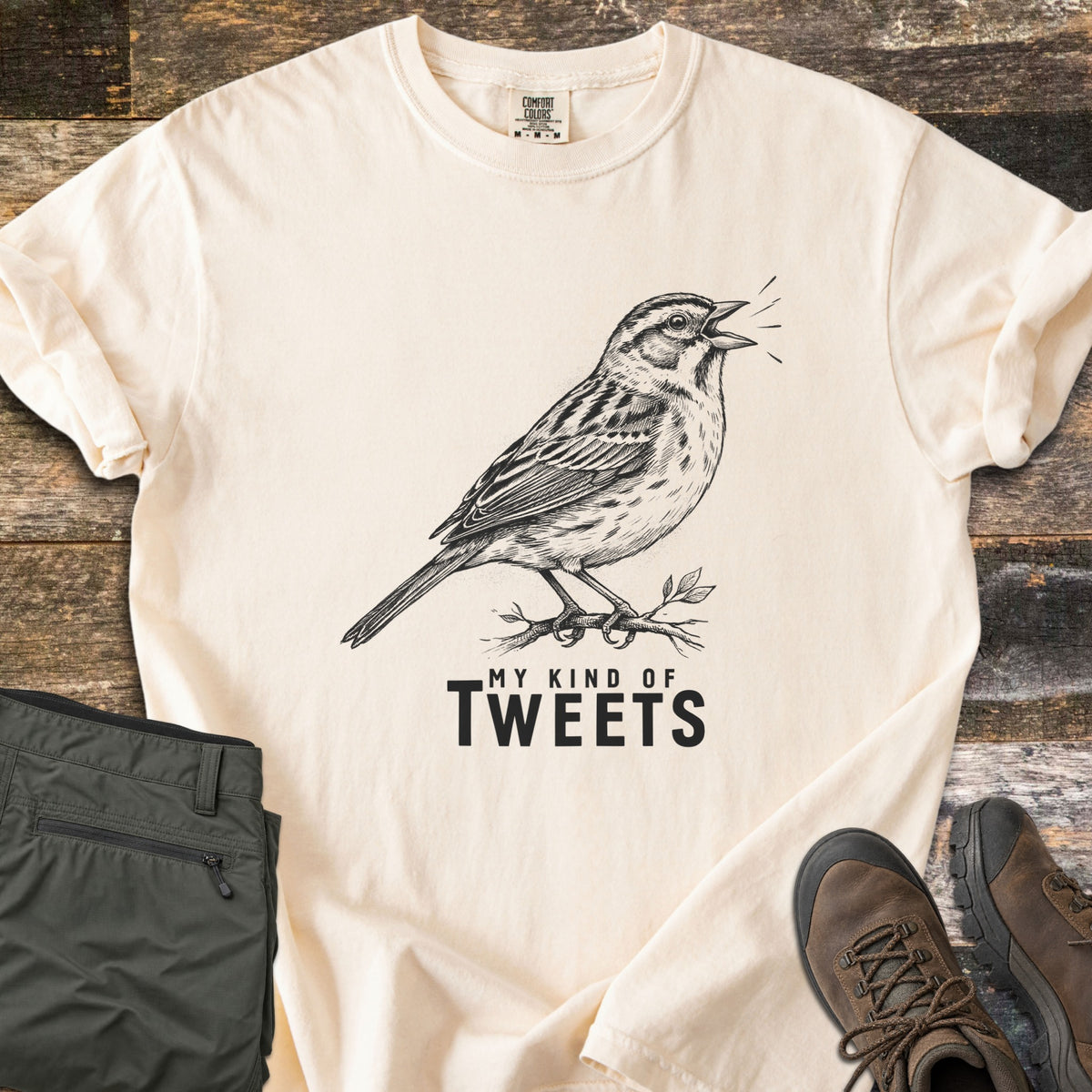My Kind Of Tweets Comfort Colors T-shirt in Ivory, laid flat. A black sketch of a singing sparrow perched on a leafy twig, above the words My Kind Of Tweets. Shown on rustic wood with folded olive hiking pants and brown hiking boots.