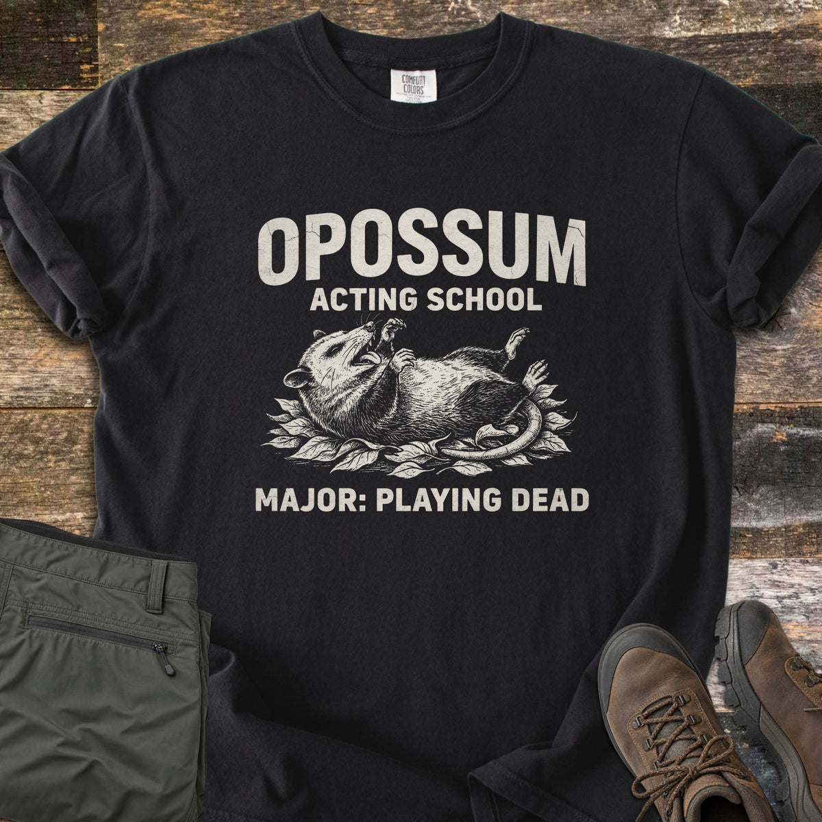 Opossum Acting School Comfort Colors T-shirt in Black, laid flat. An etched drawing of an opossum lying on its back on leaves, mouth open, with the words Opossum Acting School above and Major: Playing Dead below. Sleeves rolled on wood planks beside green pants and brown hiking boots.