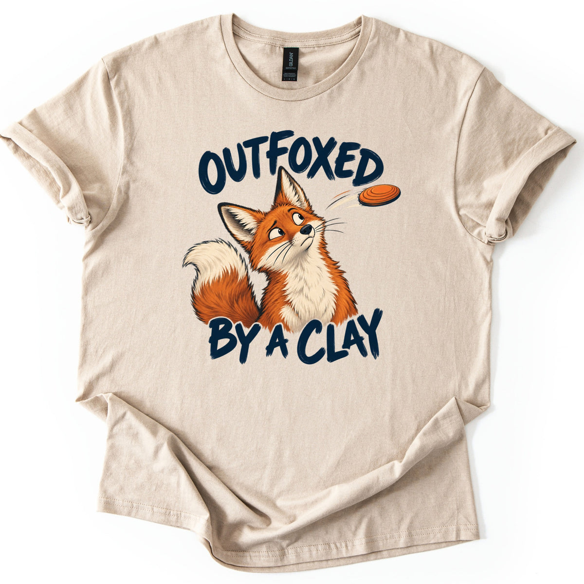 Outfoxed by a Clay Comfort Colors T-shirt in Sand, laid flat. A cartoon red fox looks up warily at an orange clay target flying past, between the dark navy brush letters OUTFOXED and BY A CLAY. The shirt has rolled sleeves and lies on a white background.