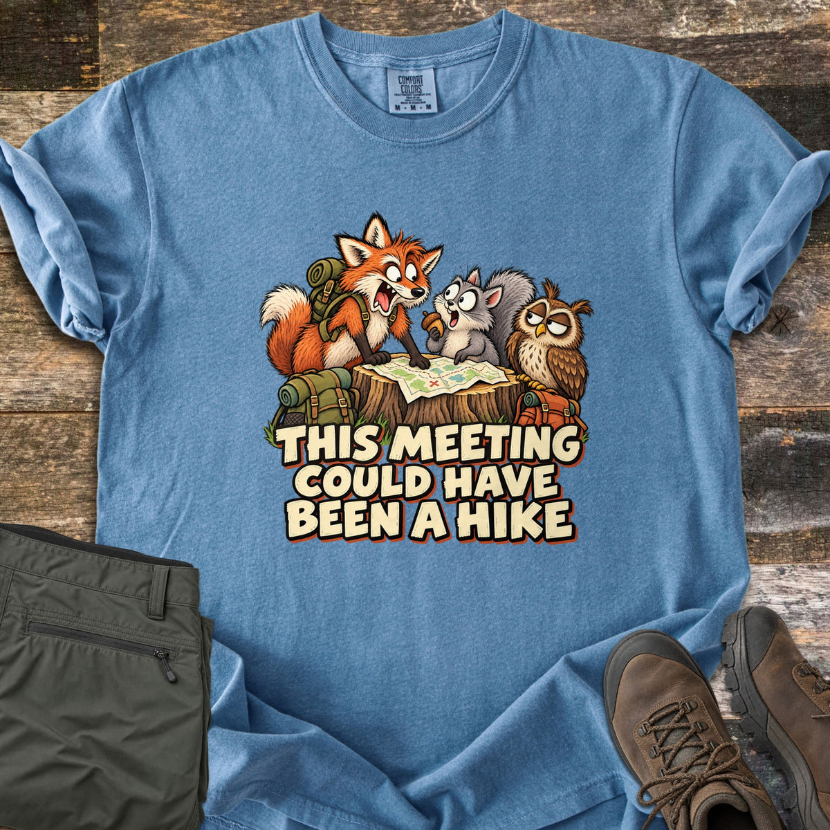 This Meeting Could Have Been a Hike Comfort Colors T-shirt in Blue Jean, laid flat. A yelling cartoon fox, a squirrel and an owl huddle over a map on a tree stump with backpacks around them, above the words This Meeting Could Have Been a Hike. Olive pants and brown boots lie on weathered wood.