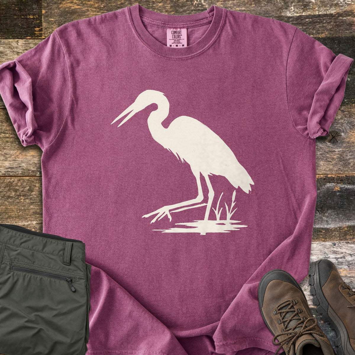 Great Blue Heron Comfort Colors T-shirt in Berry, laid flat. A cream silhouette of a heron wading with its beak open, one foot raised, beside cattails over a patch of water. Sleeves rolled, on weathered wood with olive hiking pants and brown hiking boots.