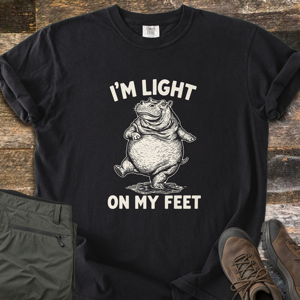I'm Light On My Feet Comfort Colors T-shirt in Black, laid flat. A sketch-style cream illustration of a round hippo dancing on one leg sits between the words I'M LIGHT and ON MY FEET. The shirt lies on weathered wood with folded olive pants and brown hiking boots.
