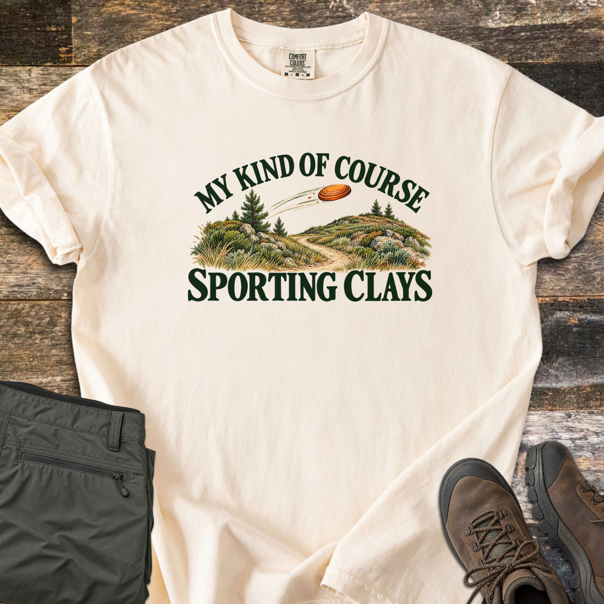 My Kind of Course Comfort Colors T-shirt in Ivory, laid flat. The words My Kind of Course arch over a painted trail winding between grassy, rocky hills with pine trees as an orange clay target flies past, above Sporting Clays. Olive pants and brown hiking boots sit on weathered wood.