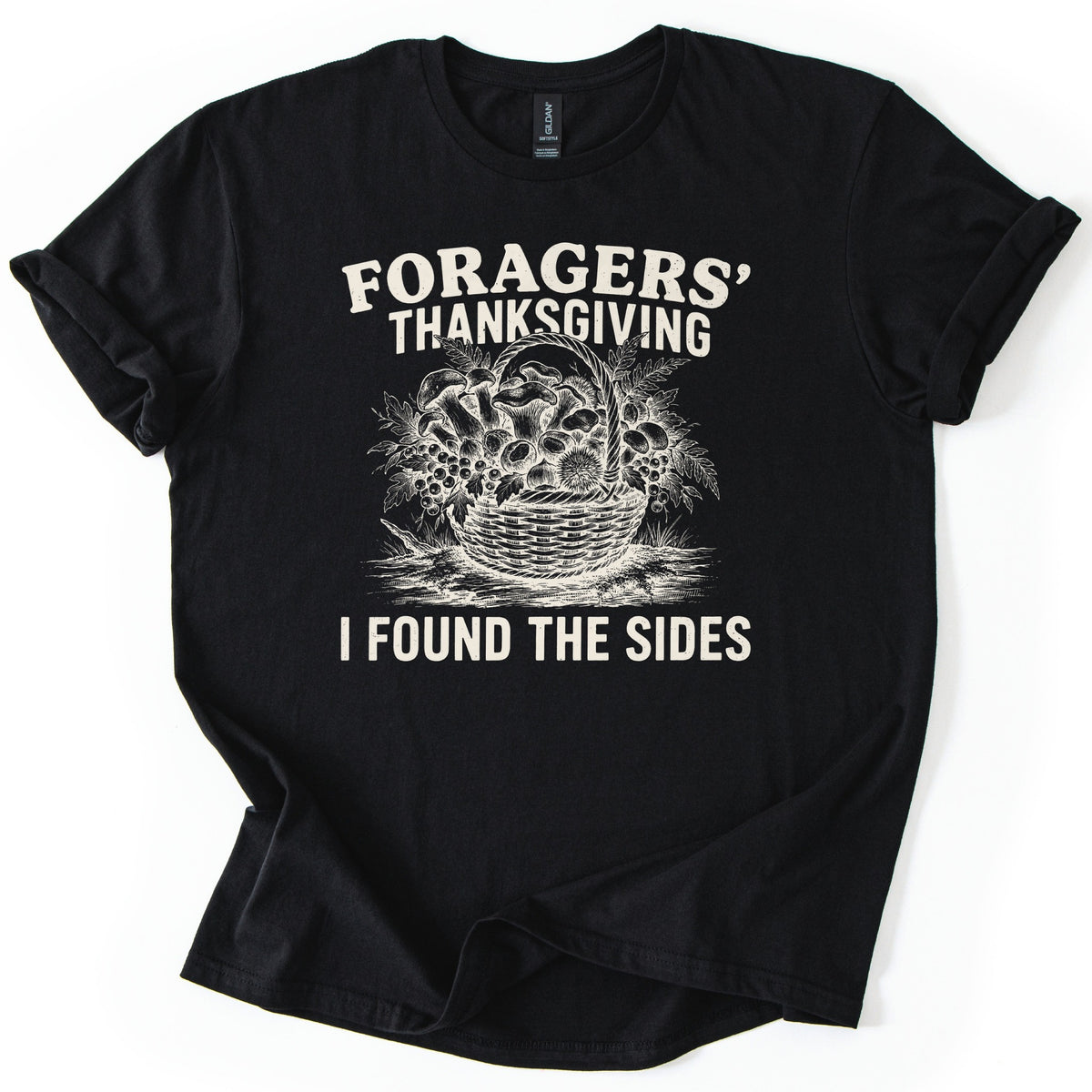 Foragers Thanksgiving Comfort Colors T-shirt in Black, laid flat. Cream sketch of a wicker basket of mushrooms, berries, ferns and a chestnut burr, between the words Foragers' Thanksgiving and I Found the Sides. Sleeves cuffed, on a white background.