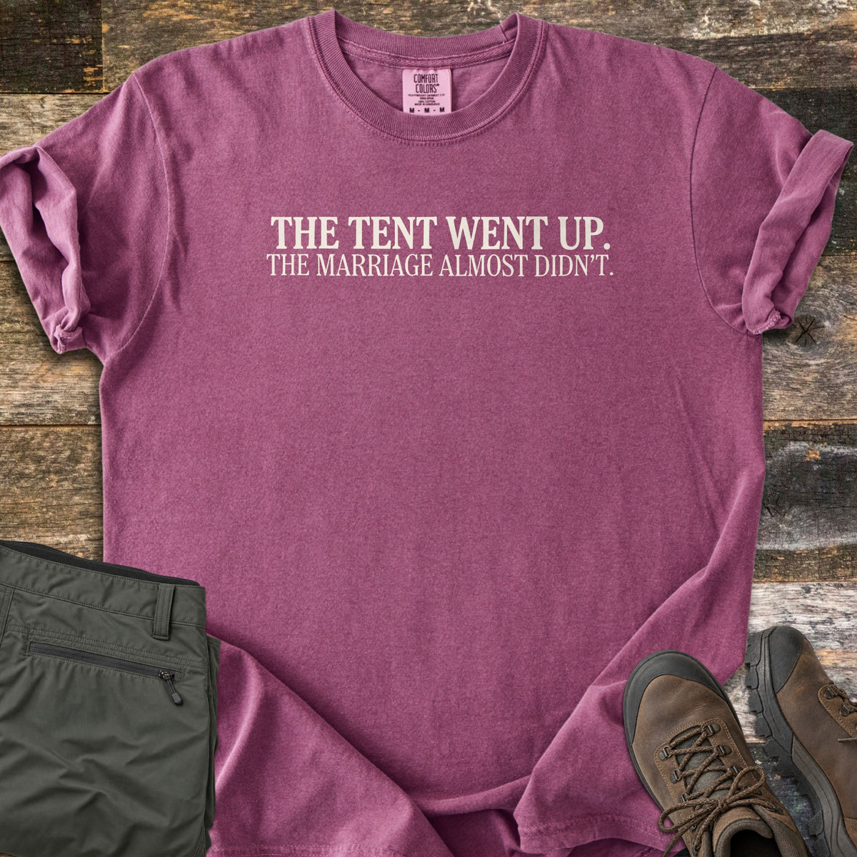 The Tent Went Up Comfort Colors T-shirt in Berry, laid flat. Cream serif text across the chest reads The Tent Went Up. The Marriage Almost Didn't. Sleeves are rolled, with folded olive hiking pants and brown hiking boots beside it on weathered wood planks.