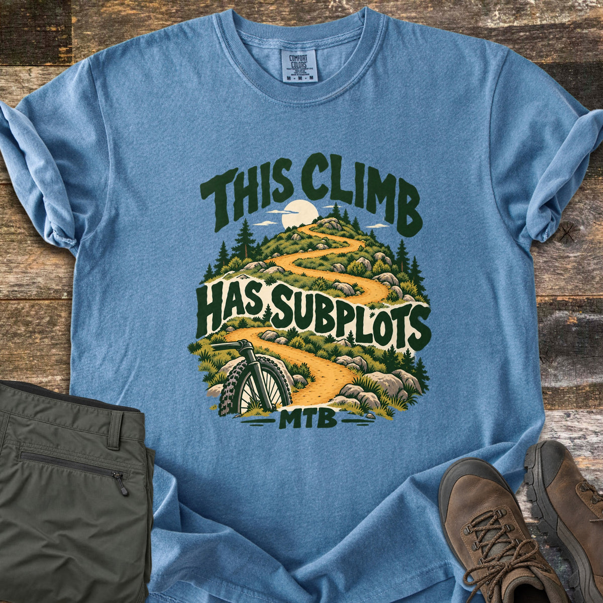 This Climb Has Subplots Comfort Colors T-shirt in Blue Jean, laid flat. A mountain bike wheel rests below a winding trail up a rocky, pine-covered hill with a pale sun, framed by the words This Climb Has Subplots MTB. Olive pants and brown hiking boots sit on weathered planks.
