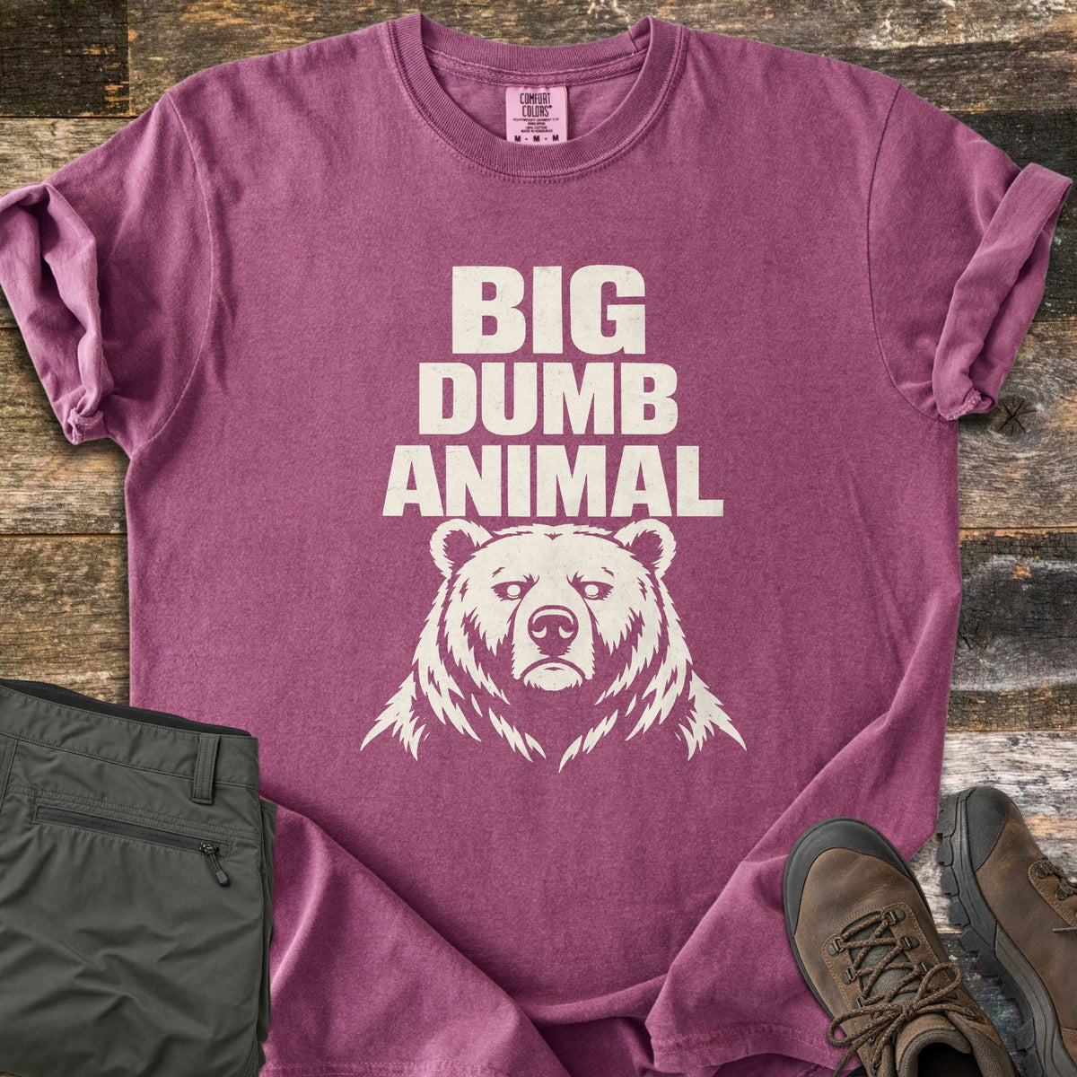 Big Dumb Animal Comfort Colors T-shirt in Berry, laid flat. Bold cream block letters read Big Dumb Animal above a grumpy-faced bear head. The shirt, sleeves rolled, lies on weathered wood planks beside folded olive hiking pants and brown hiking boots.