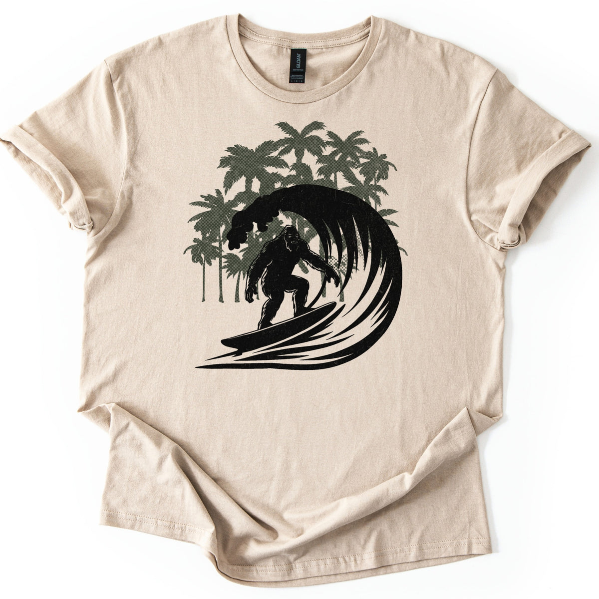 Surf Squatch Comfort Colors T-shirt in Sand, laid flat. A black silhouette of Bigfoot riding a surfboard through a curling wave, with olive green halftone palm trees behind him. No words are printed. The shirt has rolled sleeves on a white background.