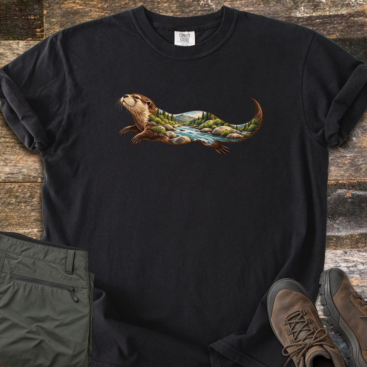 Otter River Bend Comfort Colors T-shirt in Black, laid flat. A river otter swims across the chest, its body filled with a mountain river scene of rushing water, boulders and pine trees. Olive pants and brown hiking boots lie beside it on weathered wood planks.